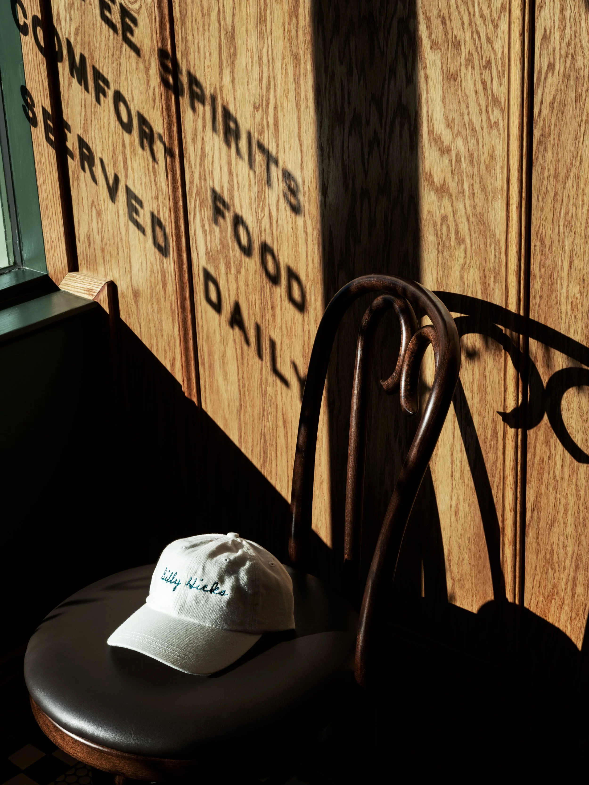 A white baseball cap with blue embroidery resting on a black leather seat, with sunlight casting shadows on a wooden wall behind, including a shadowed message saying 'WE APPRECIATE FOOD SERVED DAILY.'