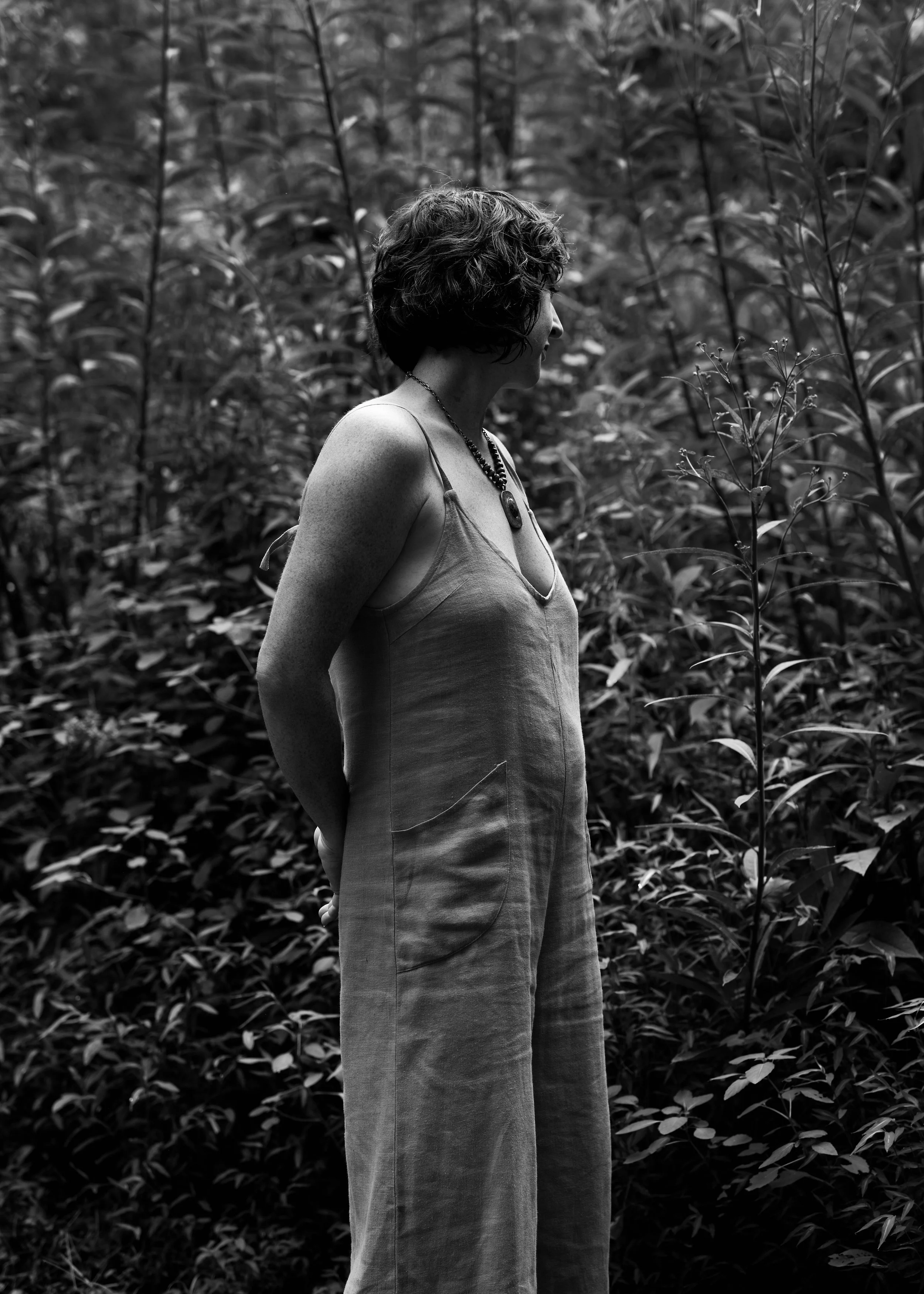 A woman standing in a forest, wearing a sleeveless top and wide-legged pants, with short curly hair, seen in profile in black and white photo.