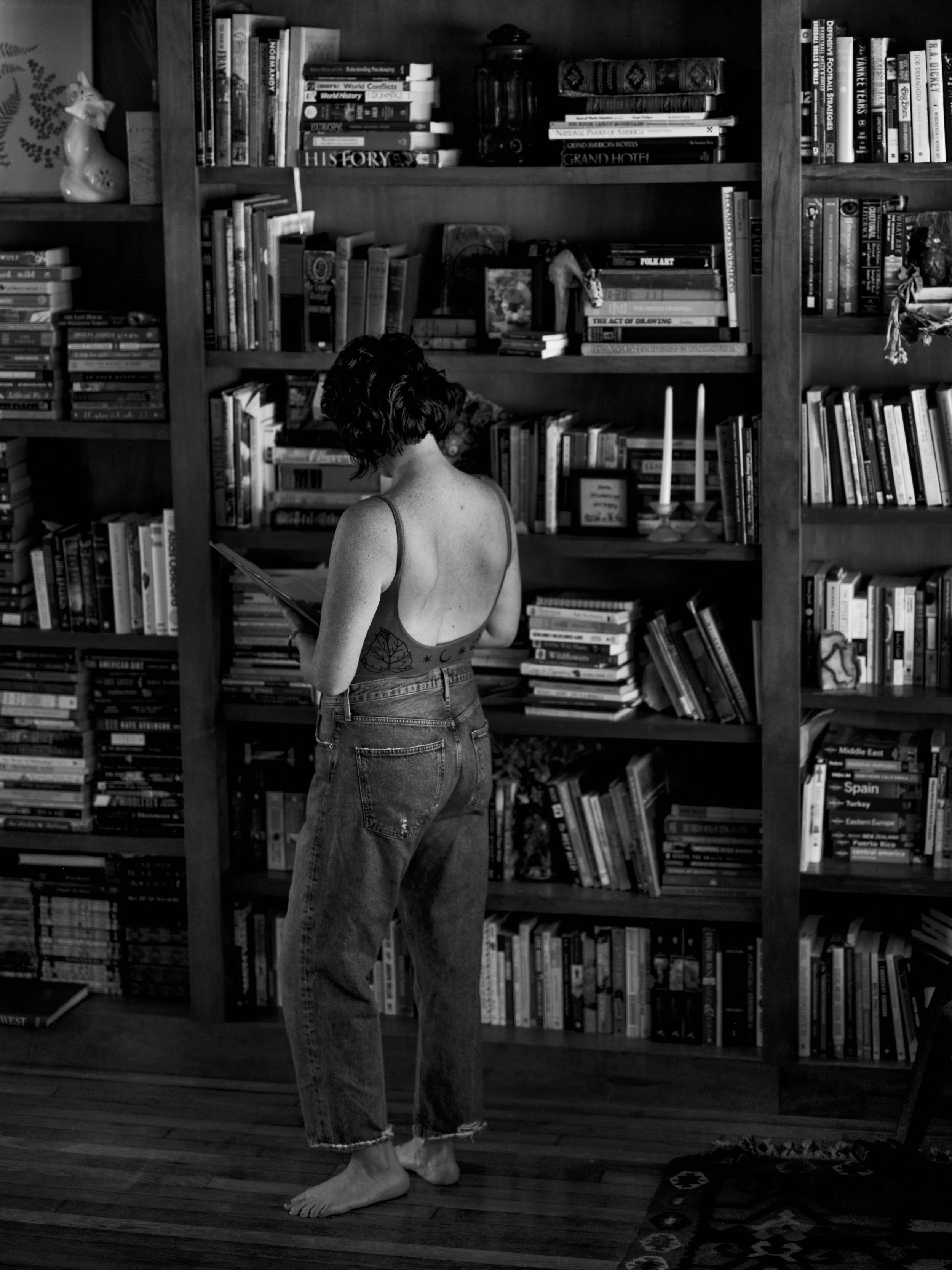 Black-and-white photo of a woman with short hair, wearing a tank top and jeans, reading a book in front of a large bookshelf filled with books, on a wooden floor.
