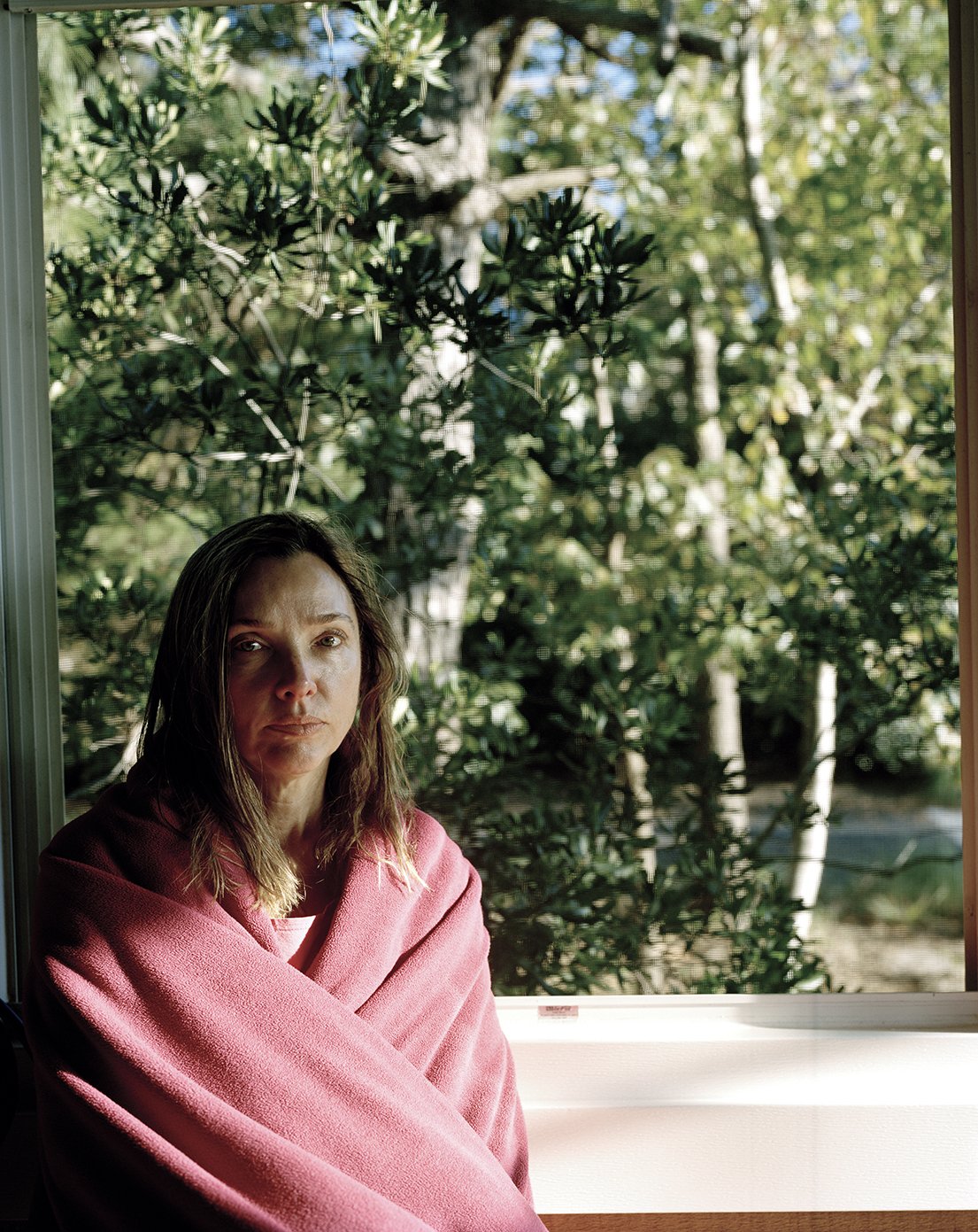 Woman wrapped in a pink blanket sitting near a window with green trees outside.