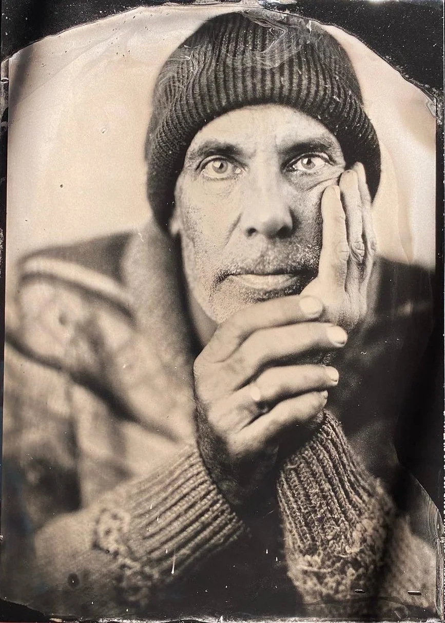 Black and white portrait of a man with a beard, wearing a beanie, holding his finger to his lips in a shushing gesture.