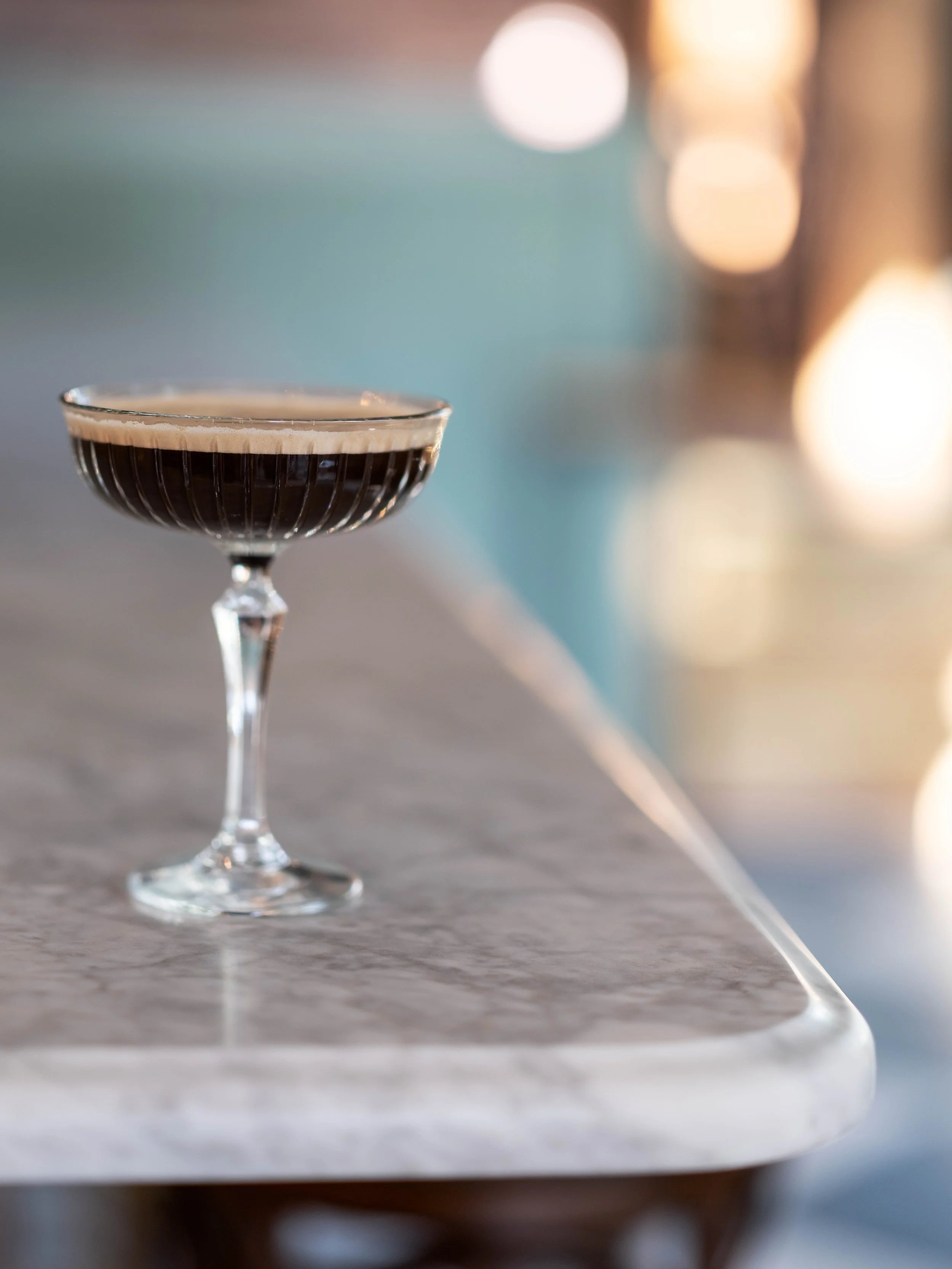 A coupe glass filled with a dark cocktail on a marble table, with blurred lights in the background.