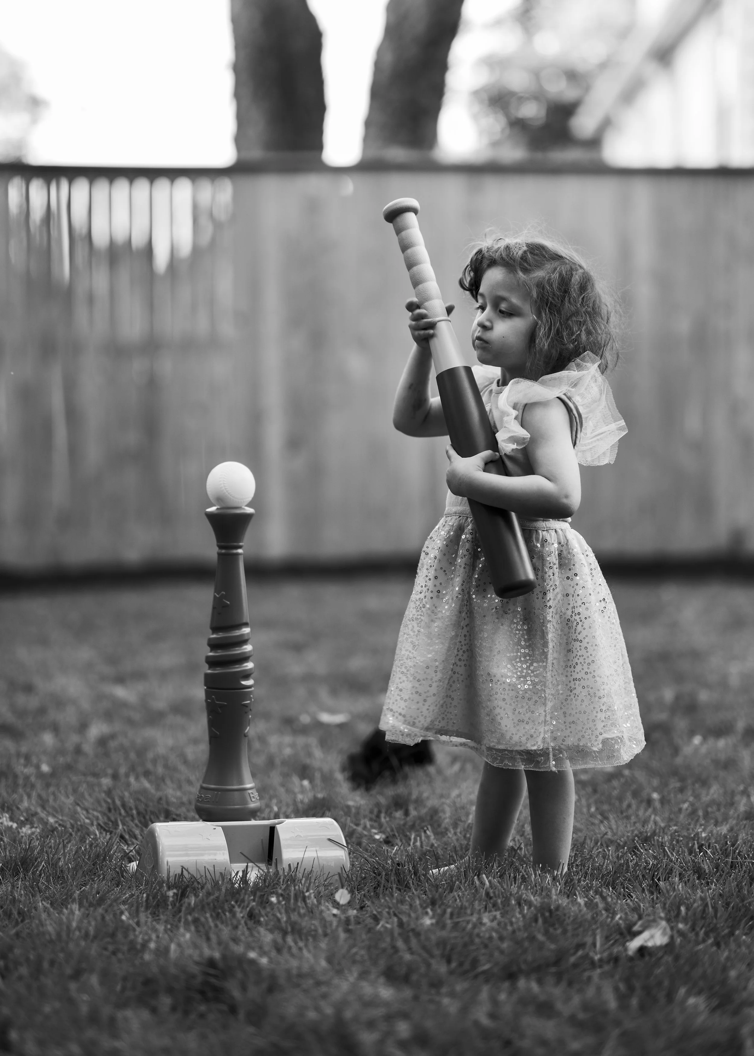 A young girl in a dress plays backyard baseball, holding a bat and preparing to hit a ball placed on a tee.