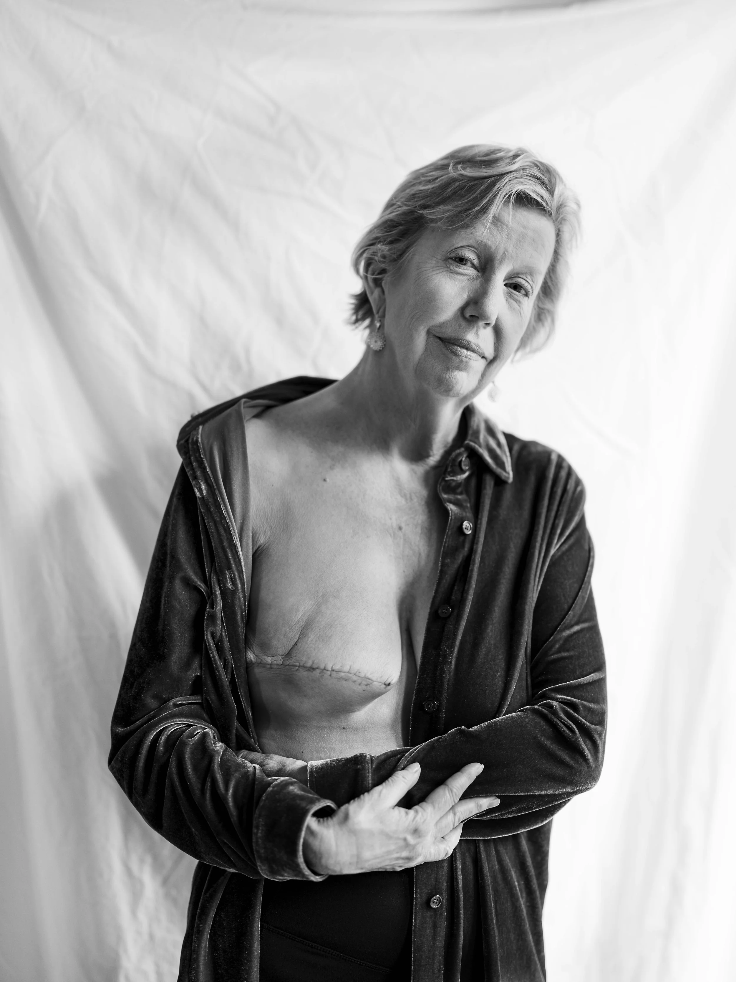 Black-and-white photo of an older woman with short, light hair smiling, wearing earrings, a partially open velvety shirt, and black pants, standing against a plain backdrop.