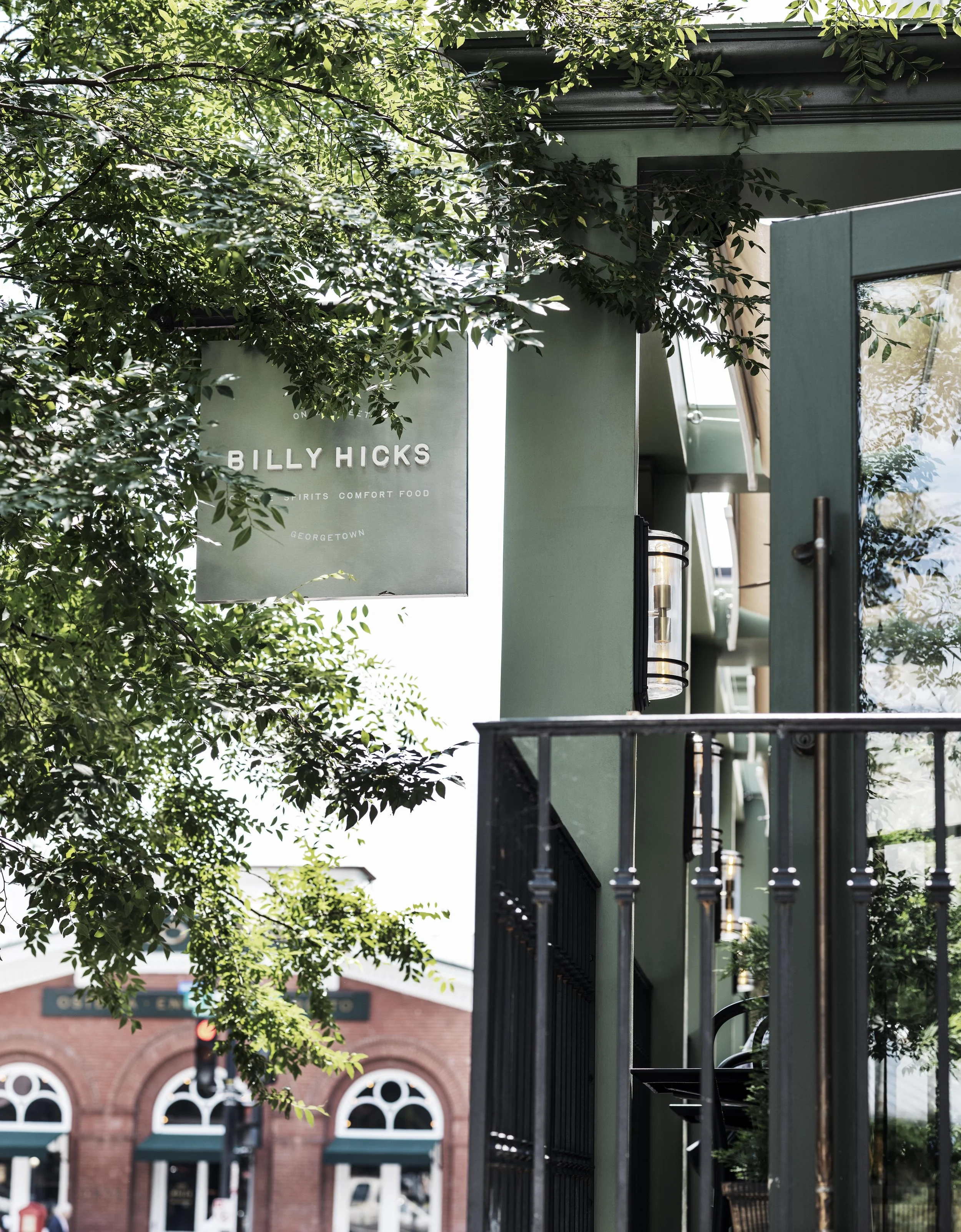 Exterior view of Billy Hicks restaurant with green facade, black metal railing, and a sign hanging from the building with the restaurant's name and description, partially obscured by tree branches.