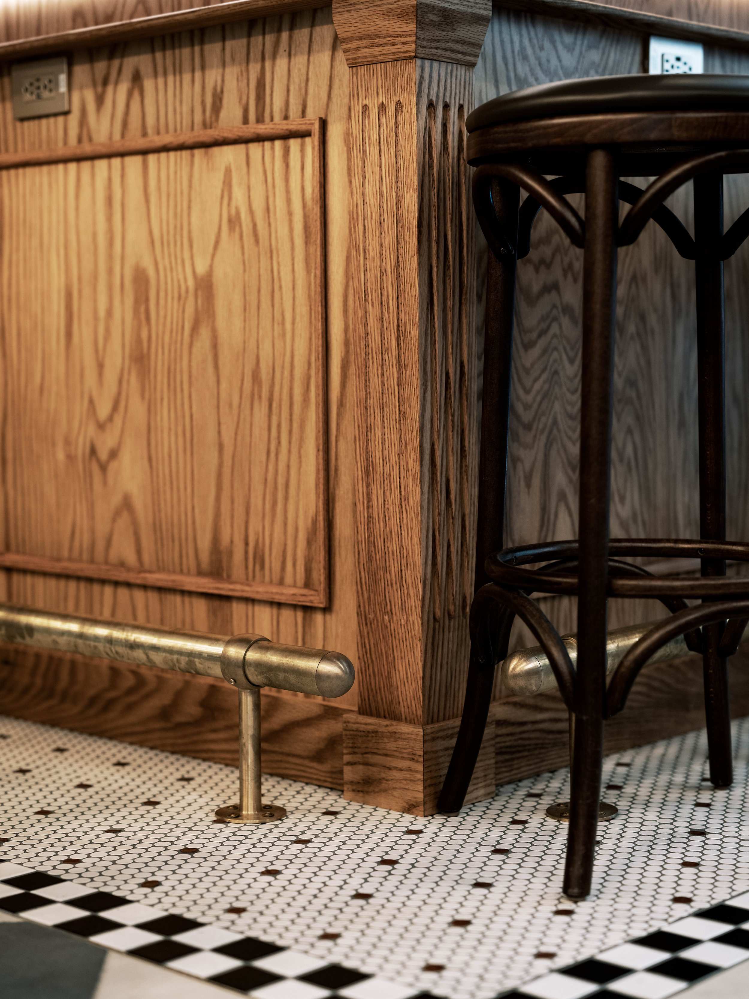 Close-up view of a wooden bar counter with a metal foot rail, a black chair, and a checkered tile floor.