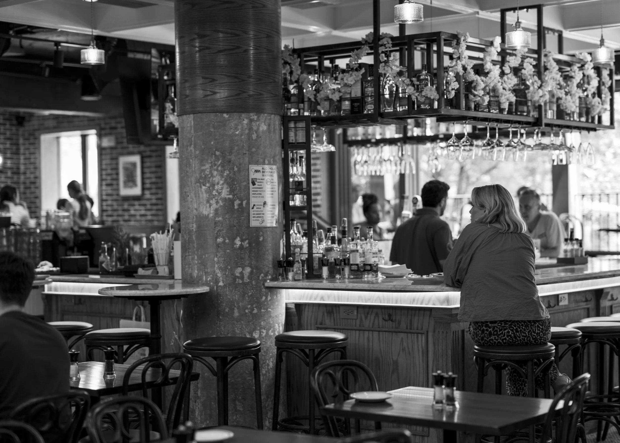 Black and white photo of a bar with a woman sitting at the counter and several patrons in the background, decorated with hanging plants and glassware.