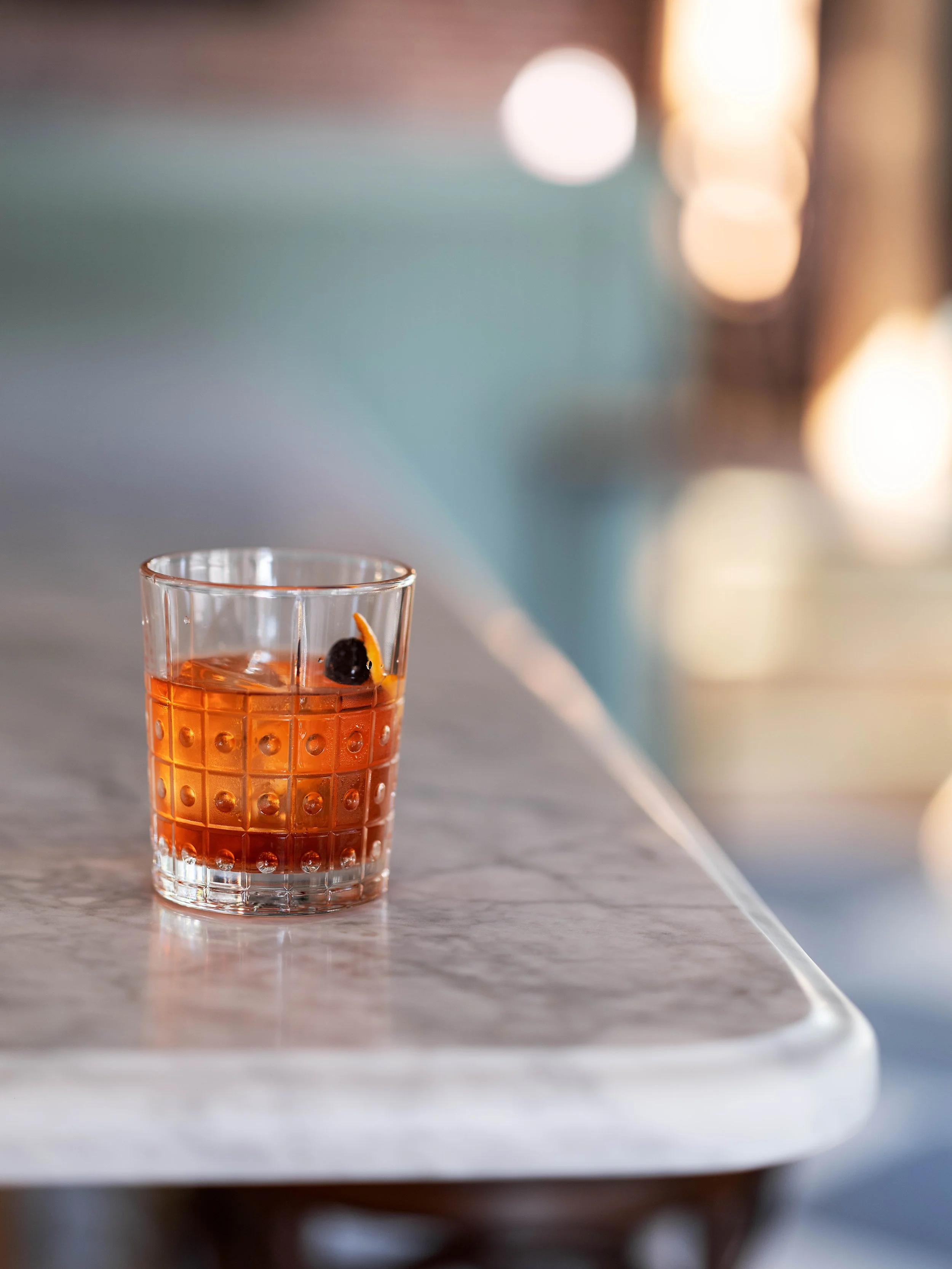 A glass of an amber-colored cocktail garnished with a cherry and an orange peel, placed on a marble table.