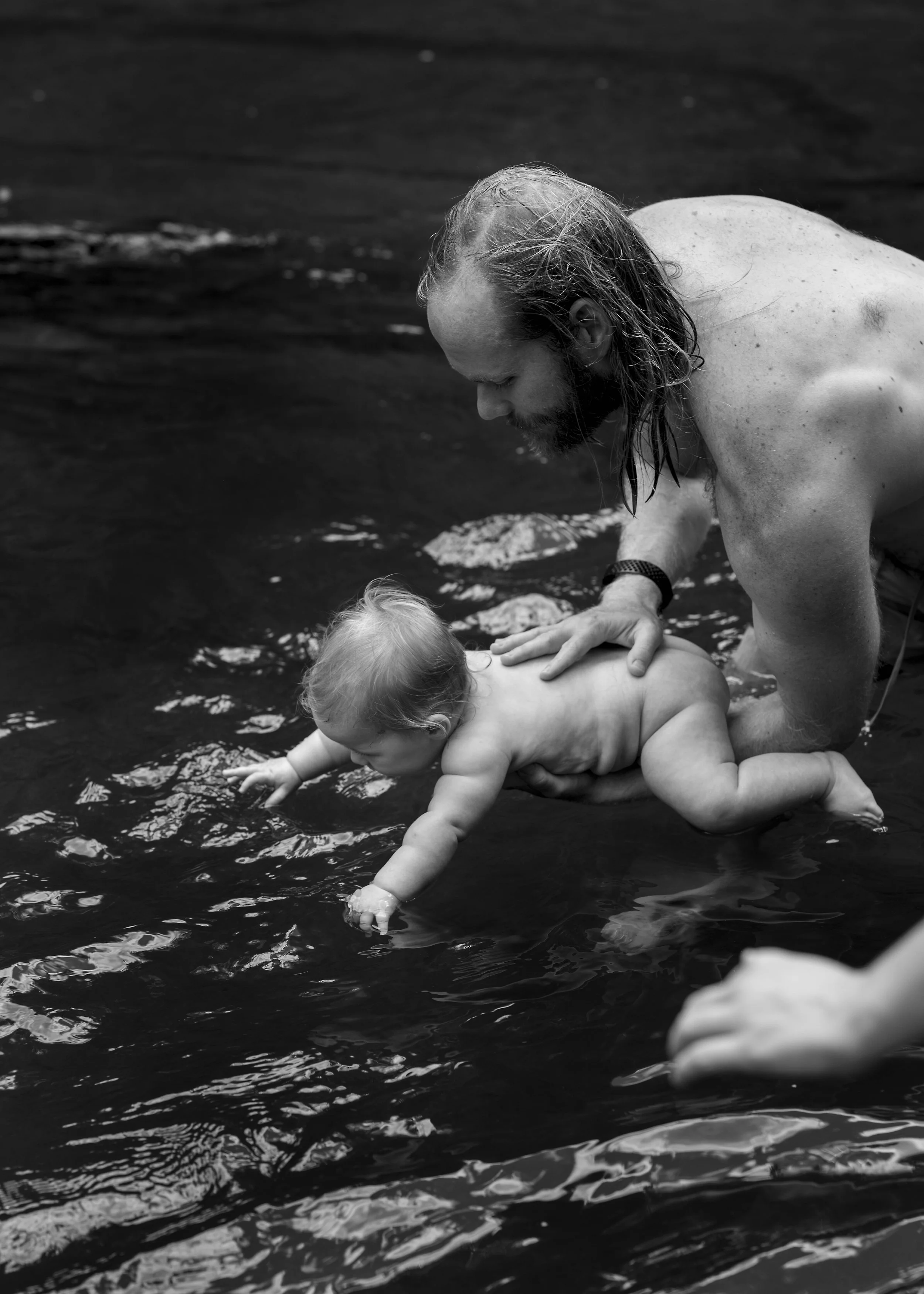 A man with long hair and a beard supports a baby as they learn to swim in a body of water. The baby is reaching into the water while the man holds them securely.
