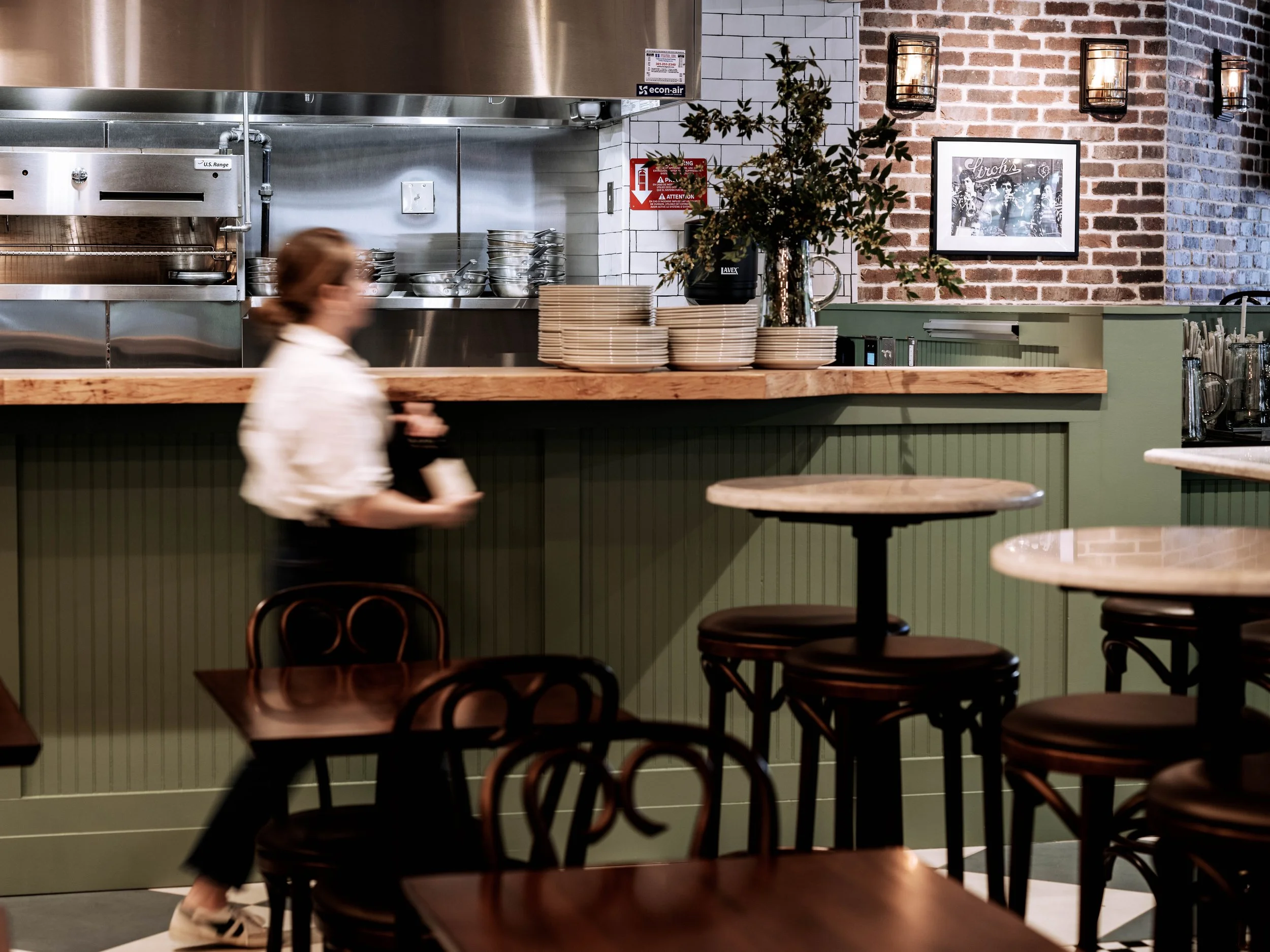 Interior of a restaurant with wooden tables and chairs, a green bar counter with stacked plates and a floral arrangement, and a blurred woman walking past.