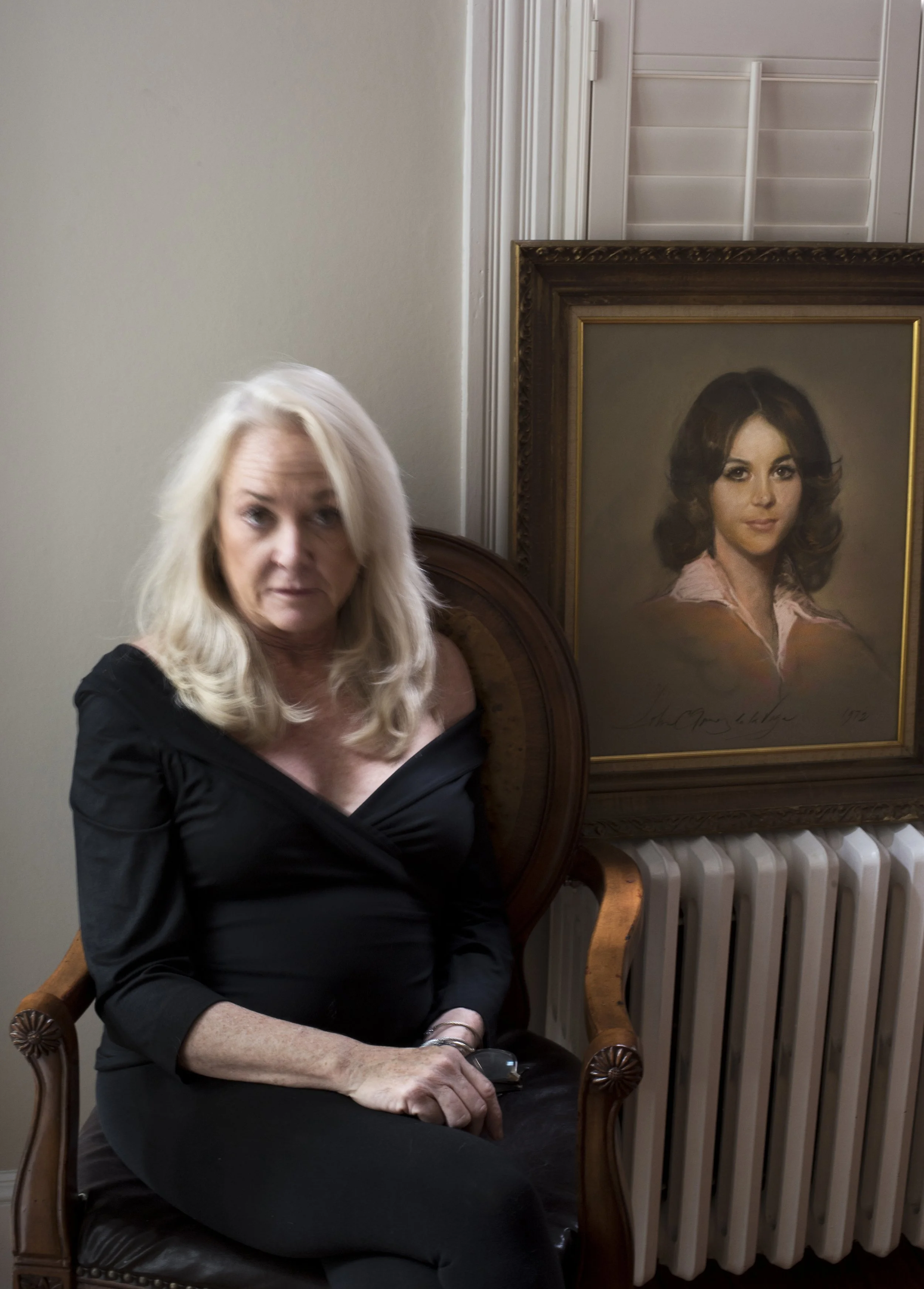 A woman with blonde hair sitting on a wooden chair with a high back, in front of a framed portrait of a woman with dark hair. The woman is wearing a black off-shoulder top and holding a small object in her right hand.
