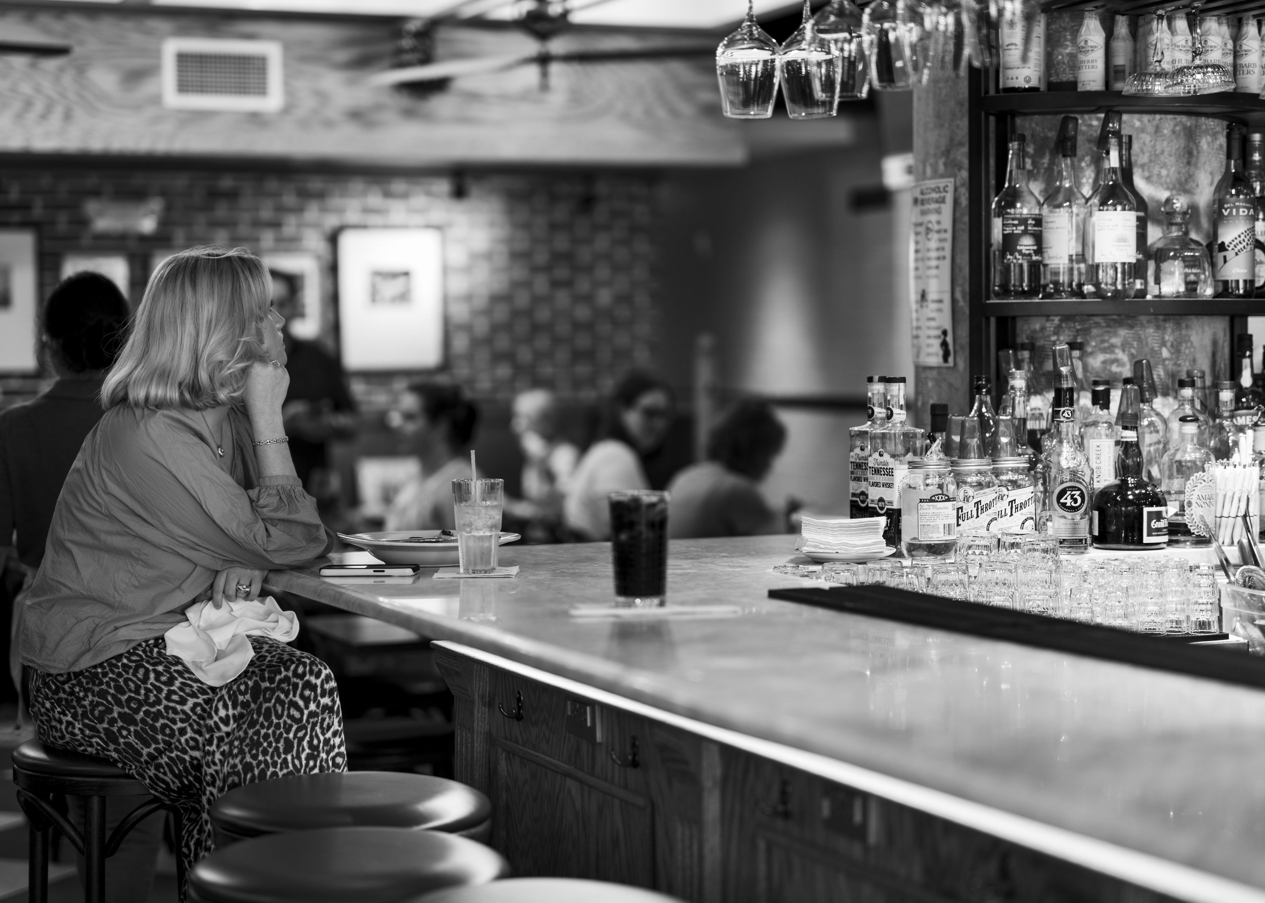 A woman sits at a bar counter in a restaurant, resting her chin on her hand, with drinks and glasses on the bar. The background shows other patrons dining and socializing.