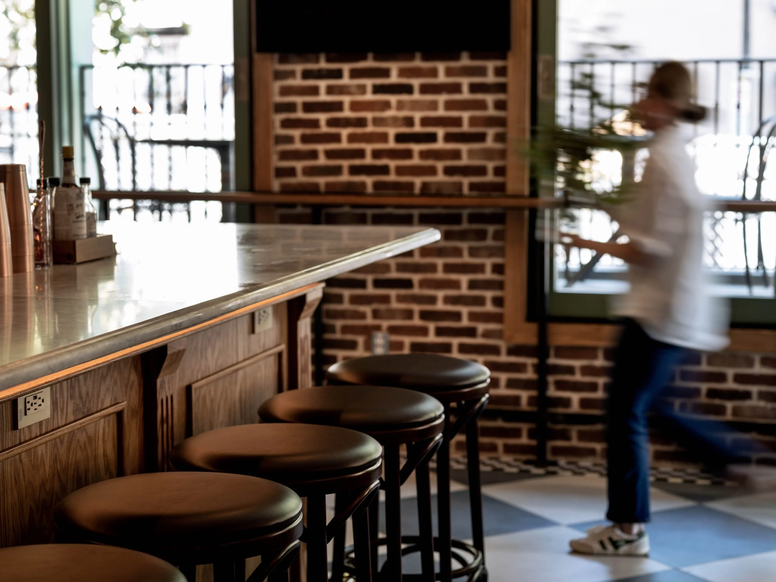 Interior of a cafe or restaurant with a counter, four brown bar stools, brick wall, and a person in a white jacket and jeans near a window, blurred due to motion.