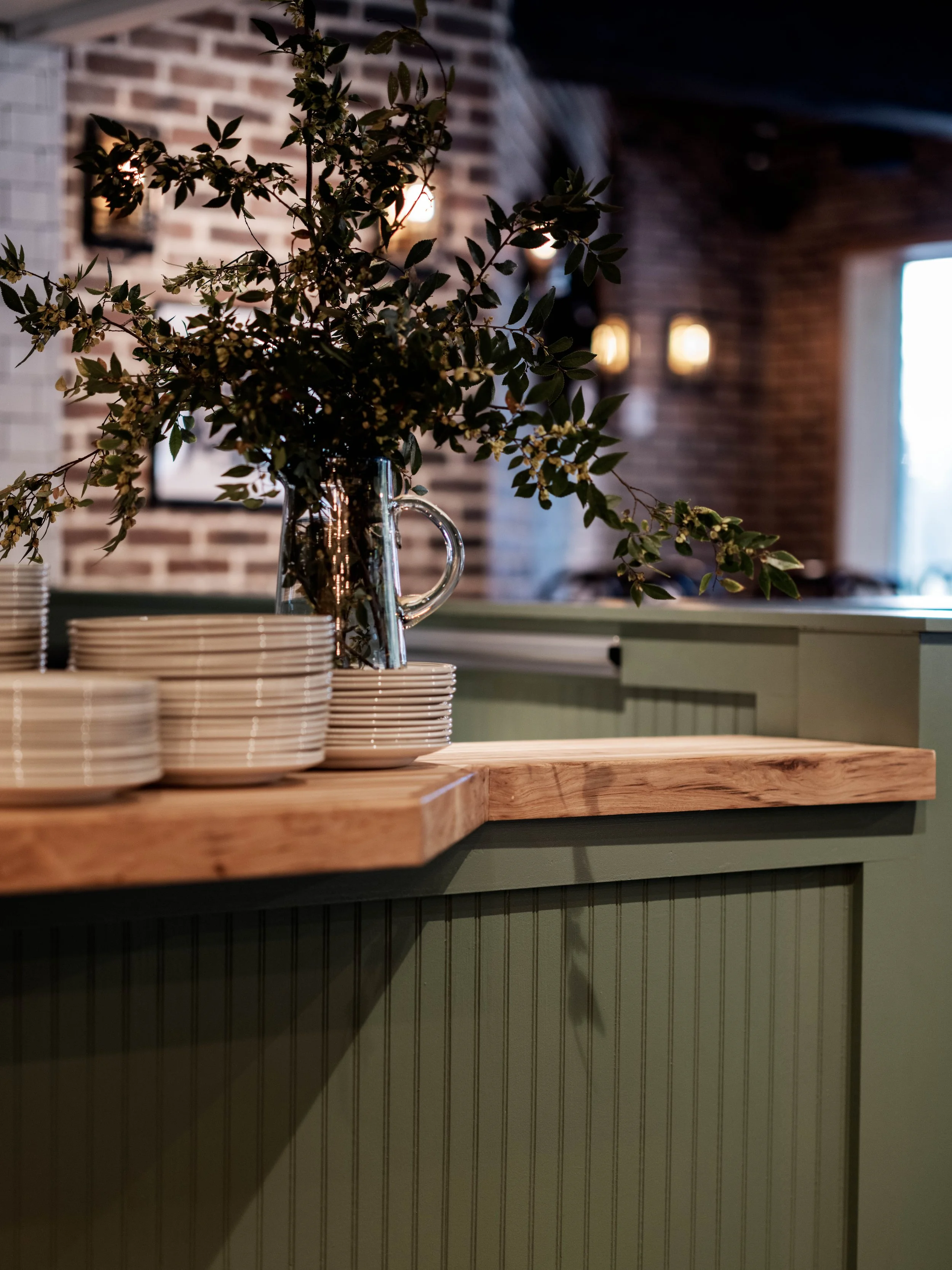 Stacked white plates and a glass pitcher with green leafy branches on a wooden counter in a rustic indoor setting.
