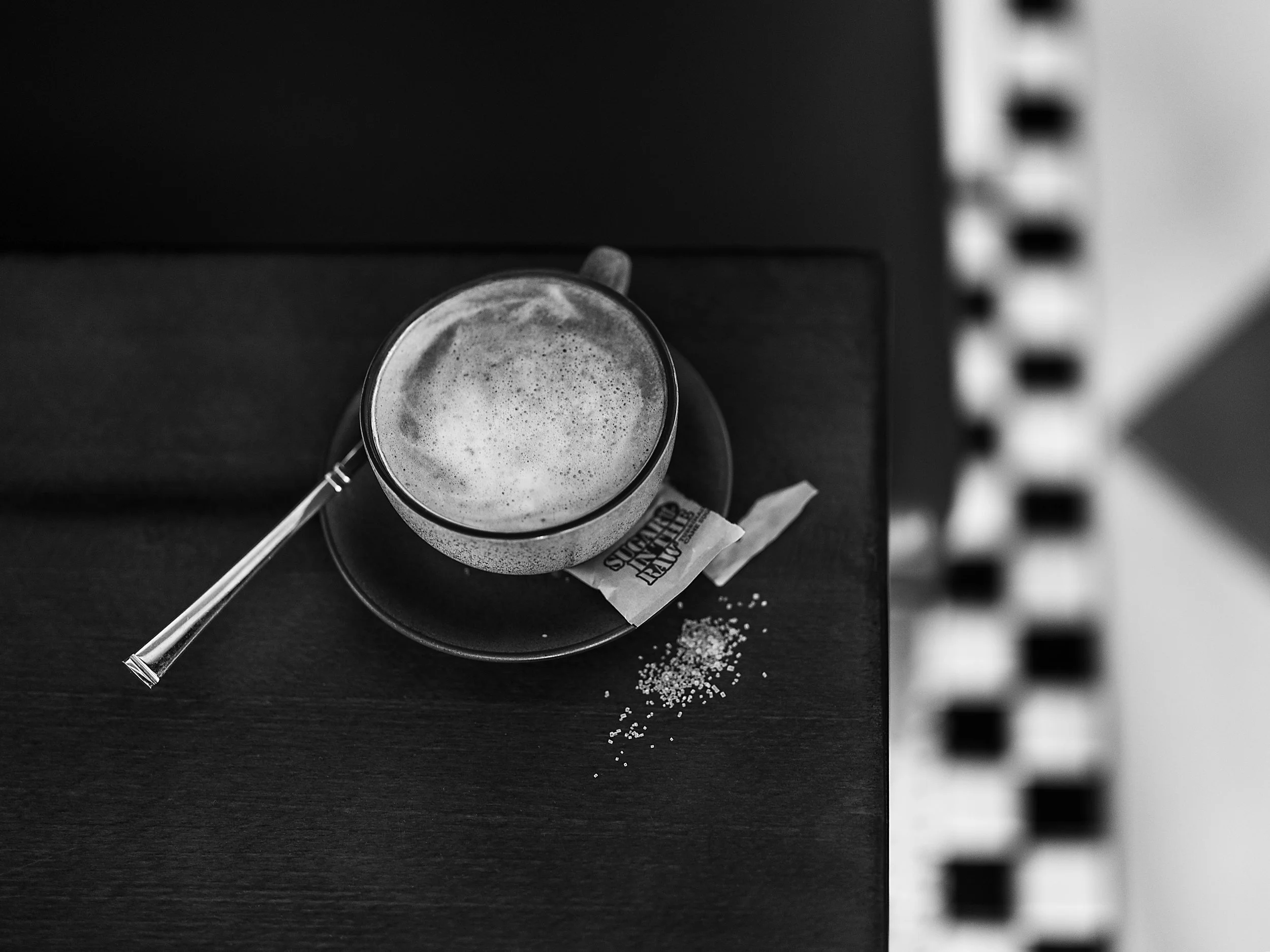 A top view black-and-white photo of a cup of frothy coffee on a saucer with a teaspoon on the side, placed on a dark table, with a sugar packet and some spilled sugar nearby.