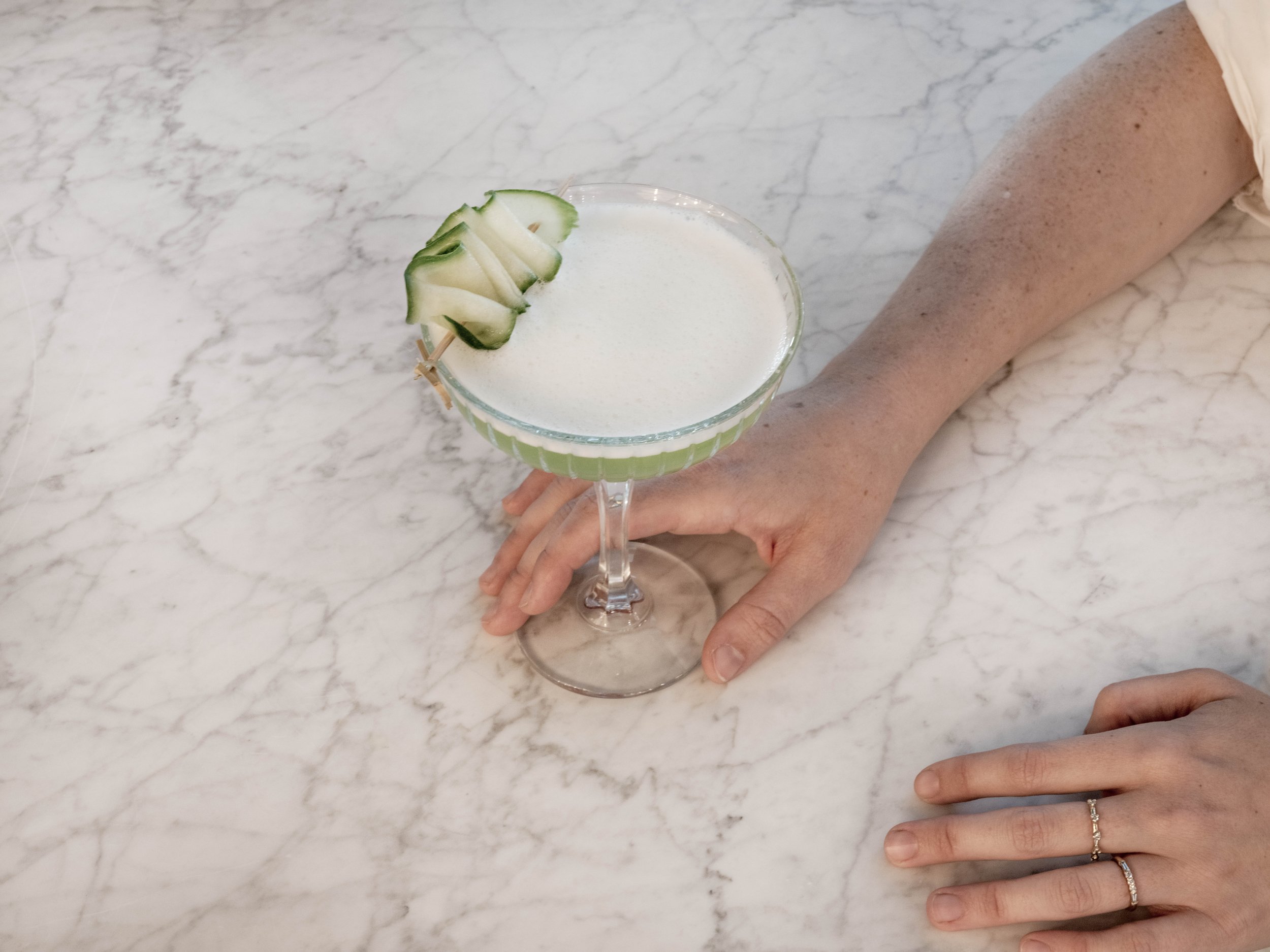 Person's hand with rings resting on a white marble surface next to a cocktail glass garnished with cucumber slices.
