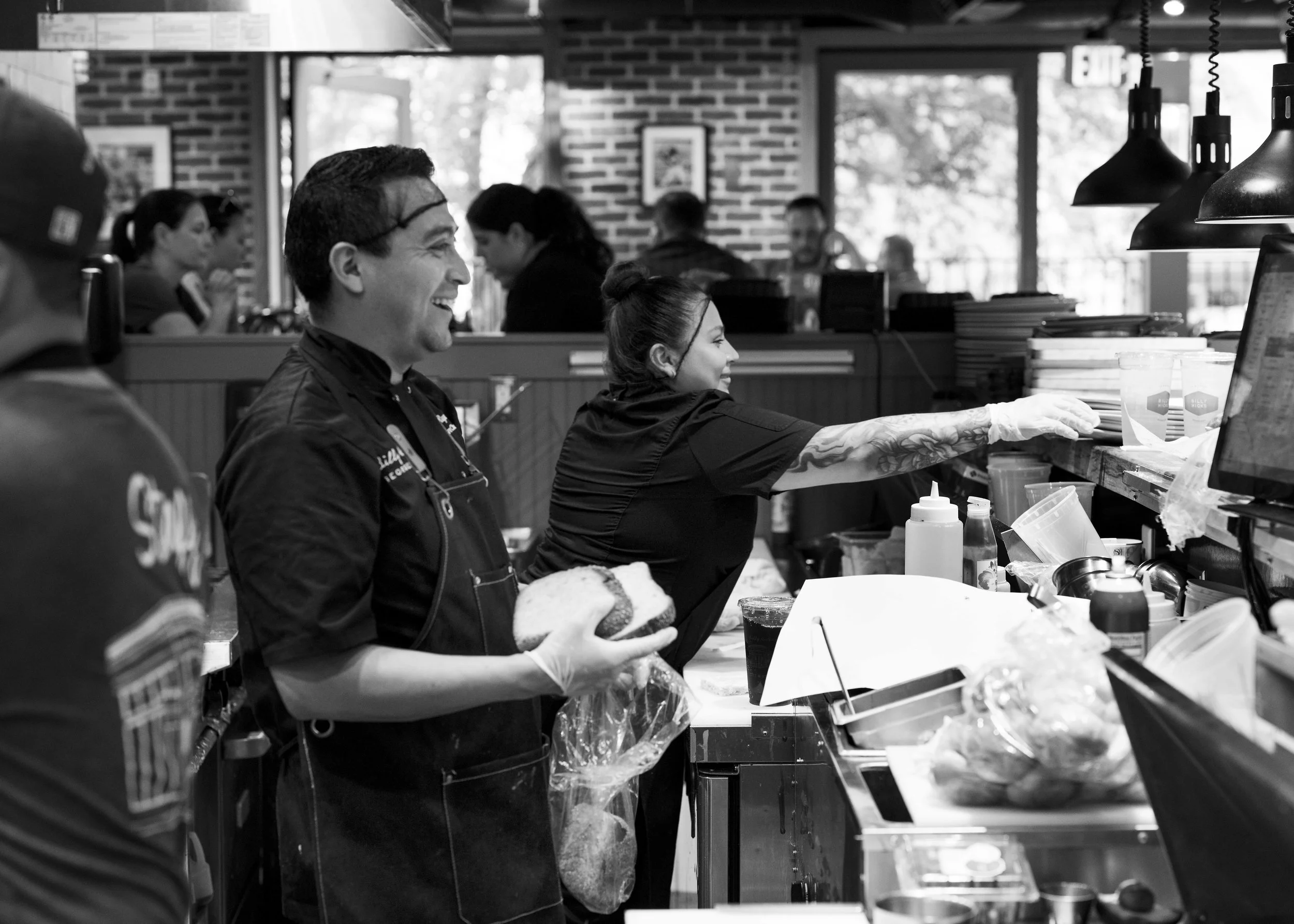 Black and white photo of a busy restaurant kitchen with smiling staff preparing food. A woman with tattoos is reaching for something at the counter, while a man in a chef's coat holds a sandwich and smiles.
