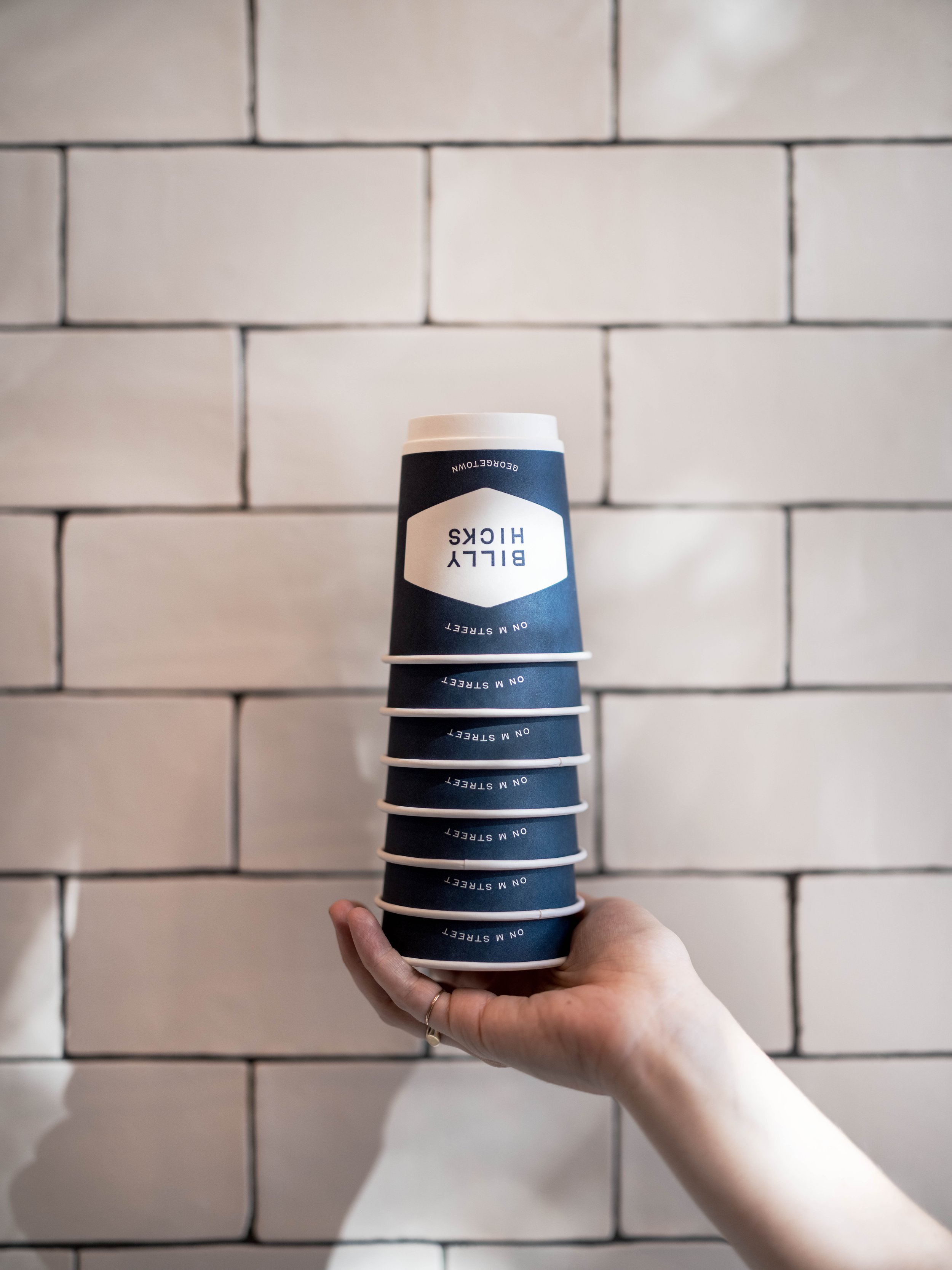 A hand holding a stack of six blue paper cups with white text and a white plastic lid on top, against a white brick wall background.