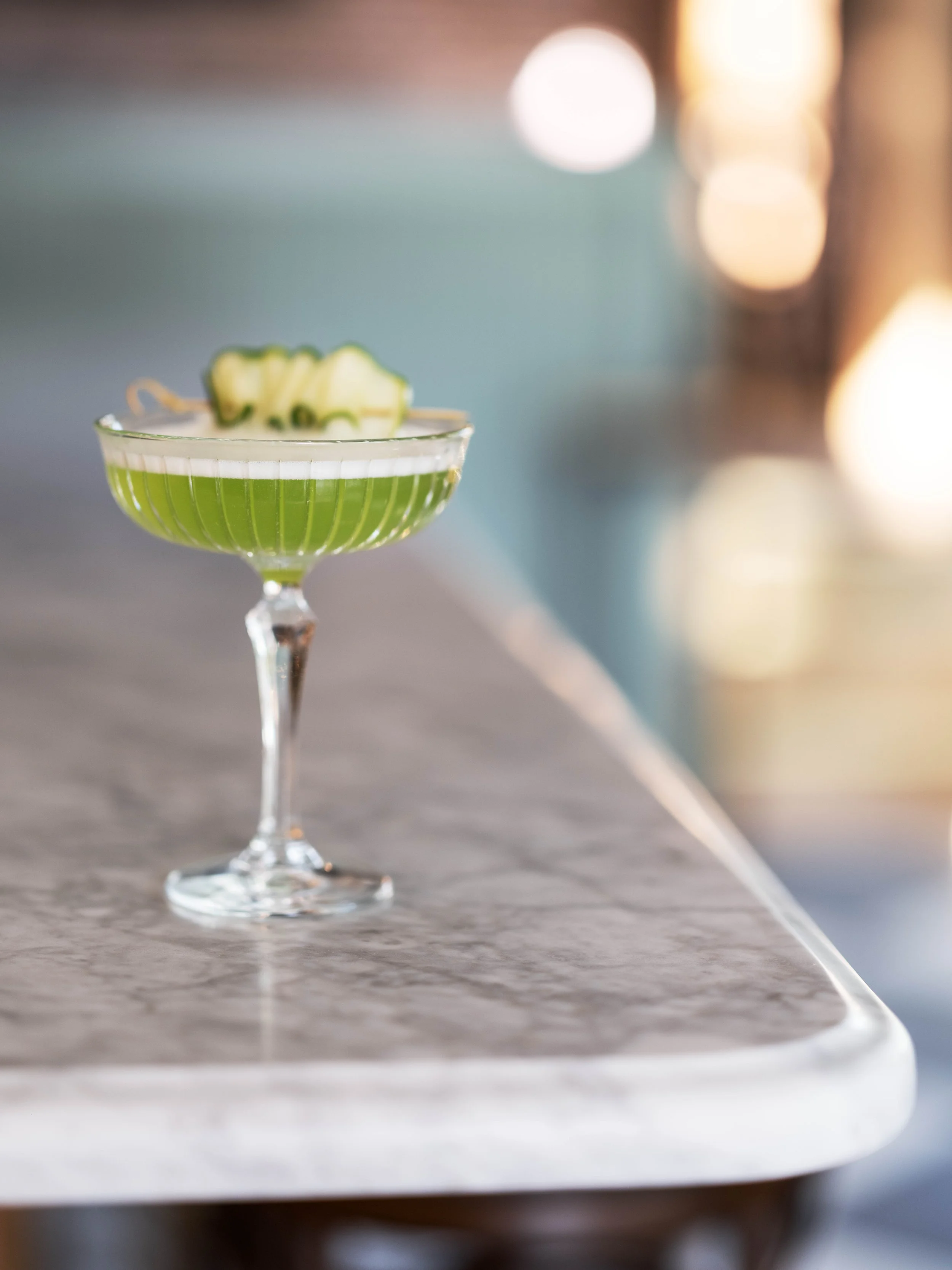 A green cocktail in a vintage-style martini glass garnished with cucumber slices, set on a marble countertop with soft blurred lights in the background.