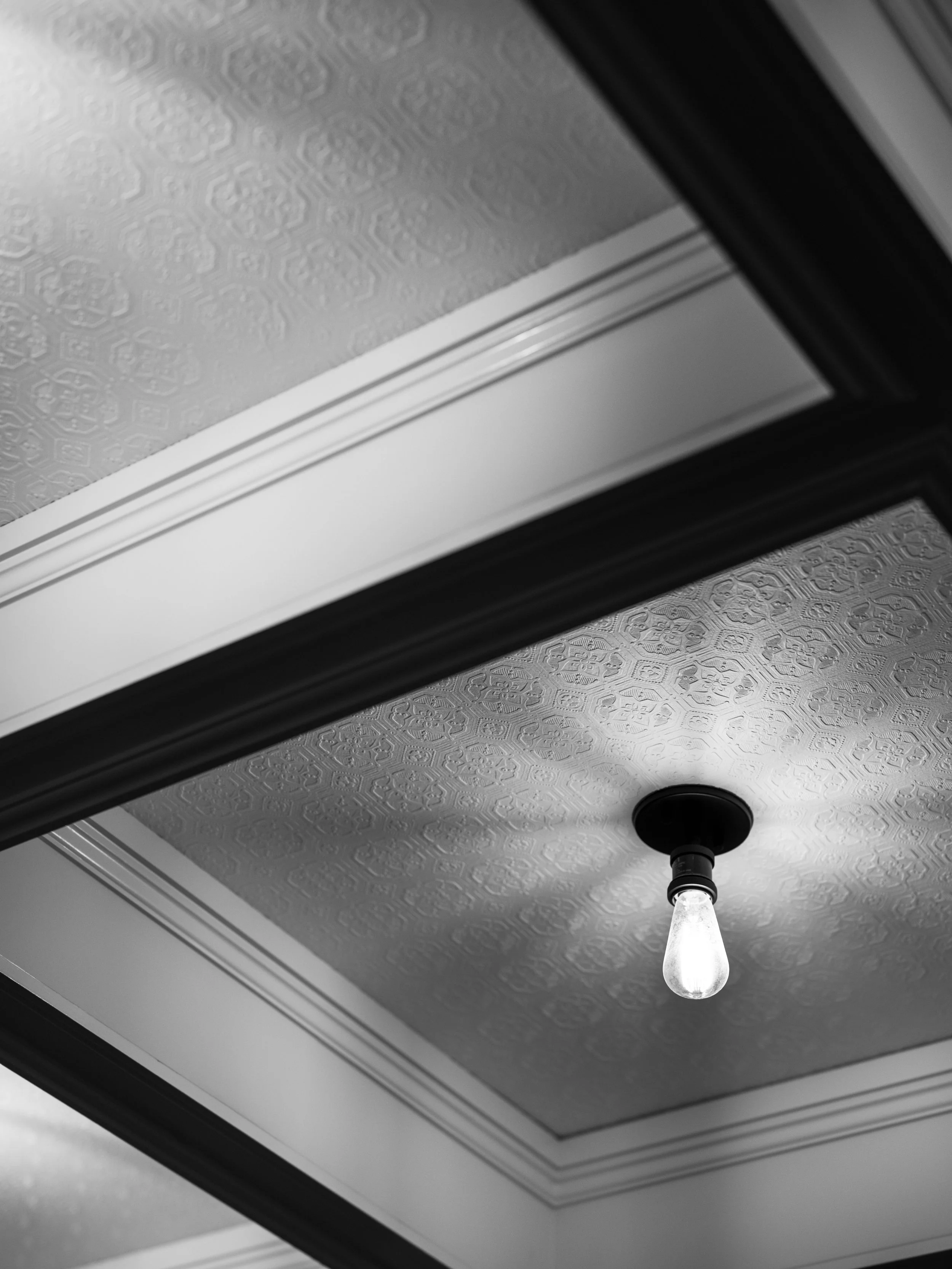 A black and white photo of a ceiling with textured wallpaper, crown molding, and a hanging light bulb fixture.