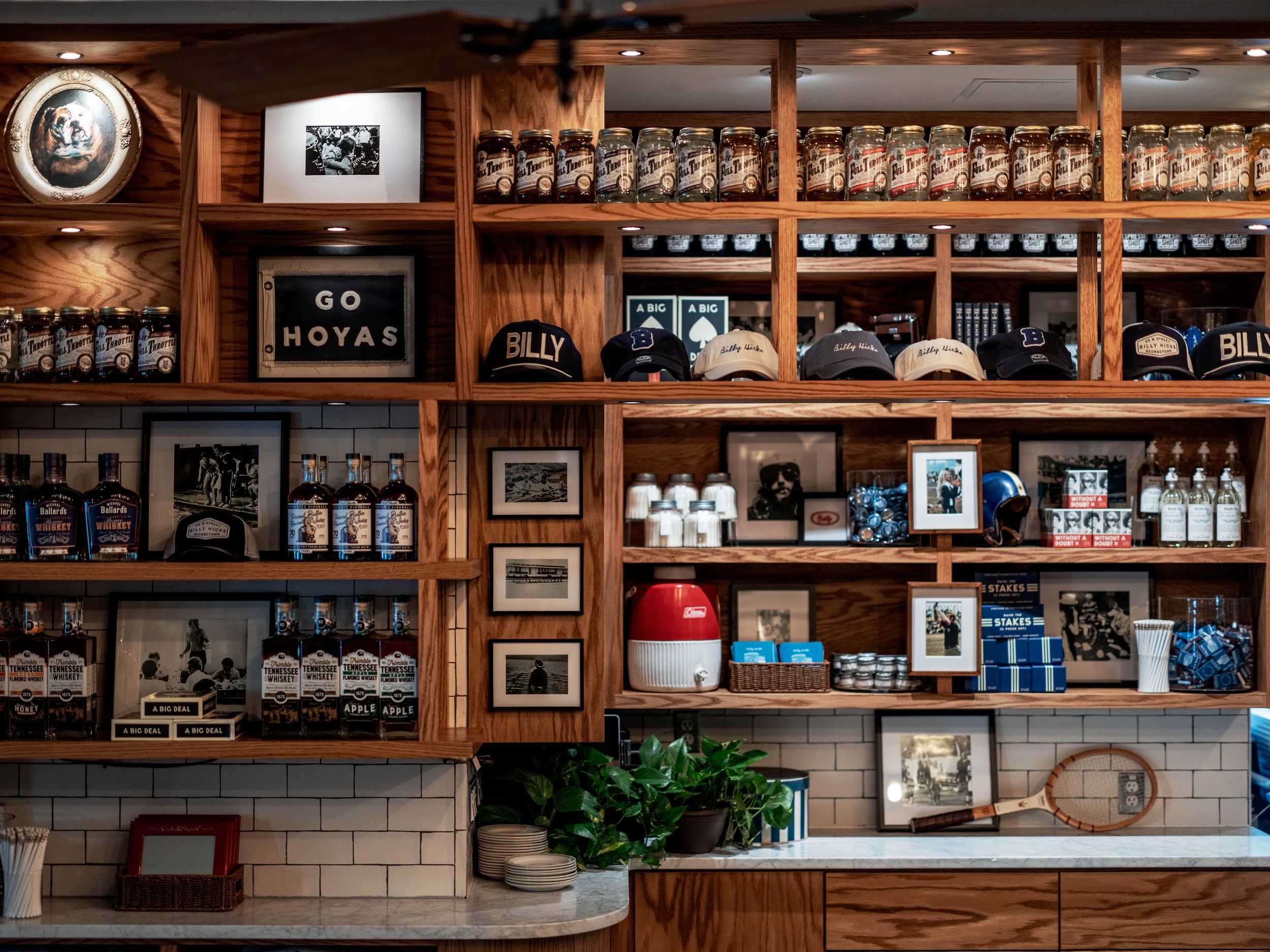 Wooden shelves with sports memorabilia, framed photos, baseball caps, jars, bottles of whiskey, a tennis racket, plants, and decorative items in a bar or restaurant interior.