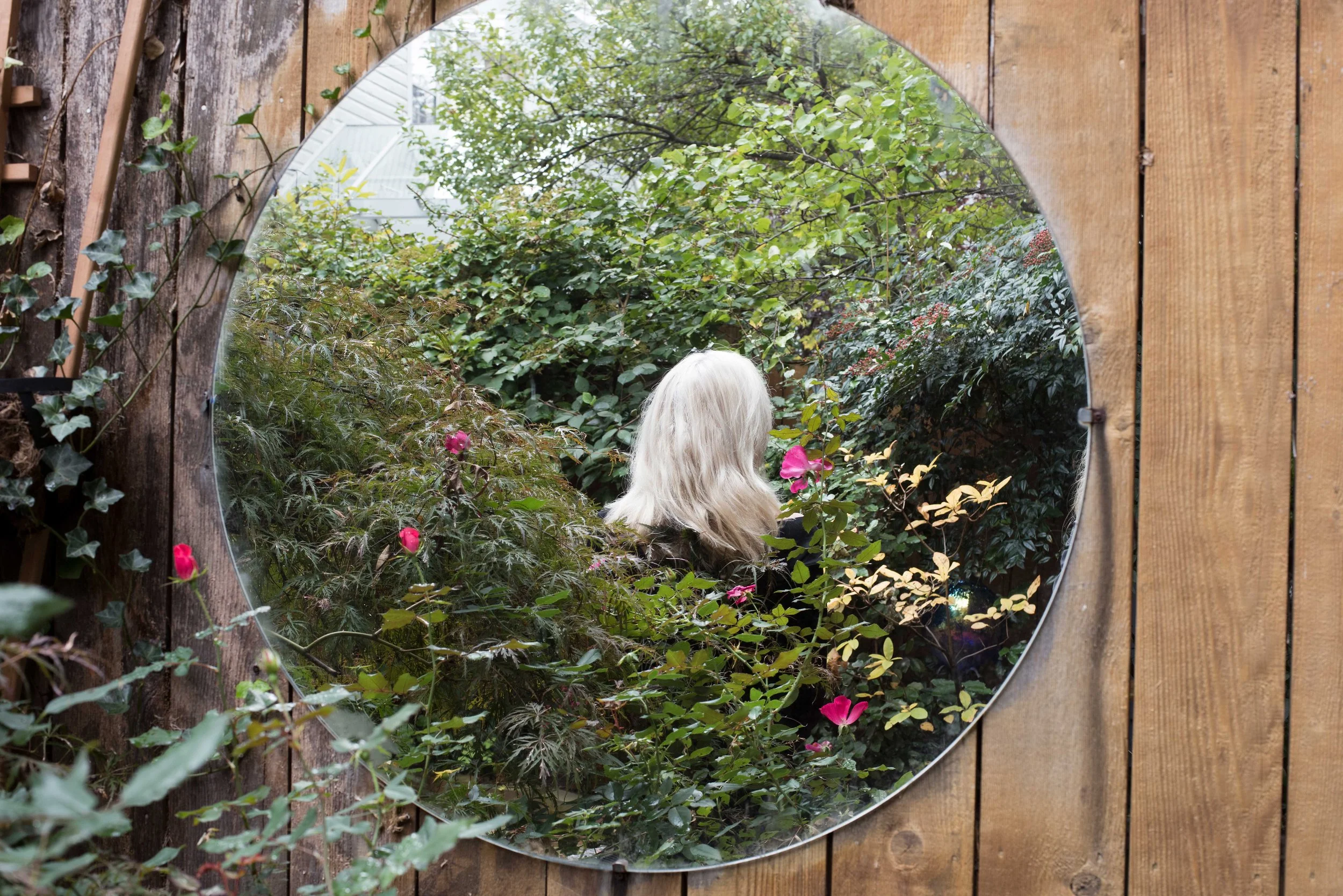 A mirror on a wooden wall reflecting a garden scene with lush green plants and pink flowers, and a woman with long white hair facing away.