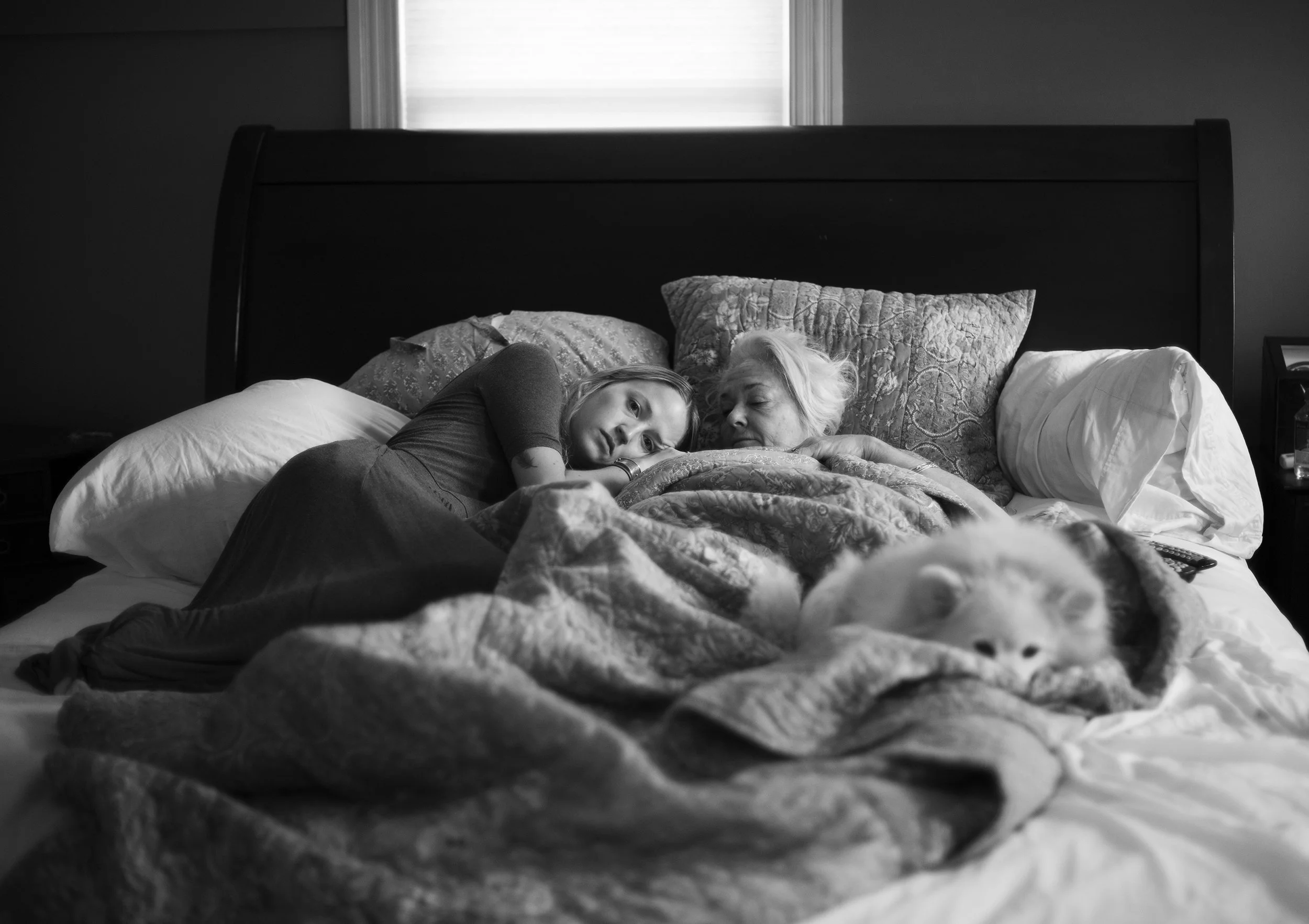 A woman and a young girl lying on a bed, resting with their heads close together, under a patterned blanket, with a fluffy dog tucked in next to them. The bed has multiple pillows, and the room has a window in the background.