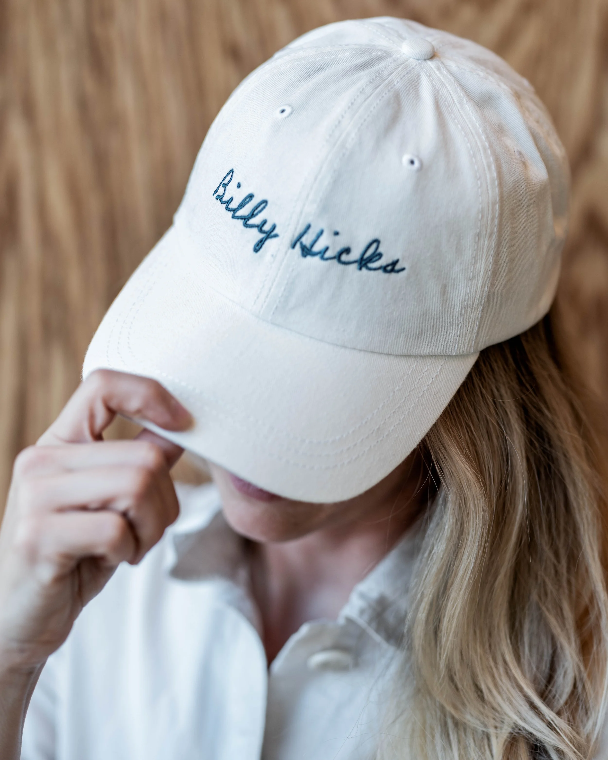 A person with long blond hair wearing a white baseball cap with the embroidered words 'Billy Kicka' on it, holding the brim of the cap with their left hand, against a wooden background.