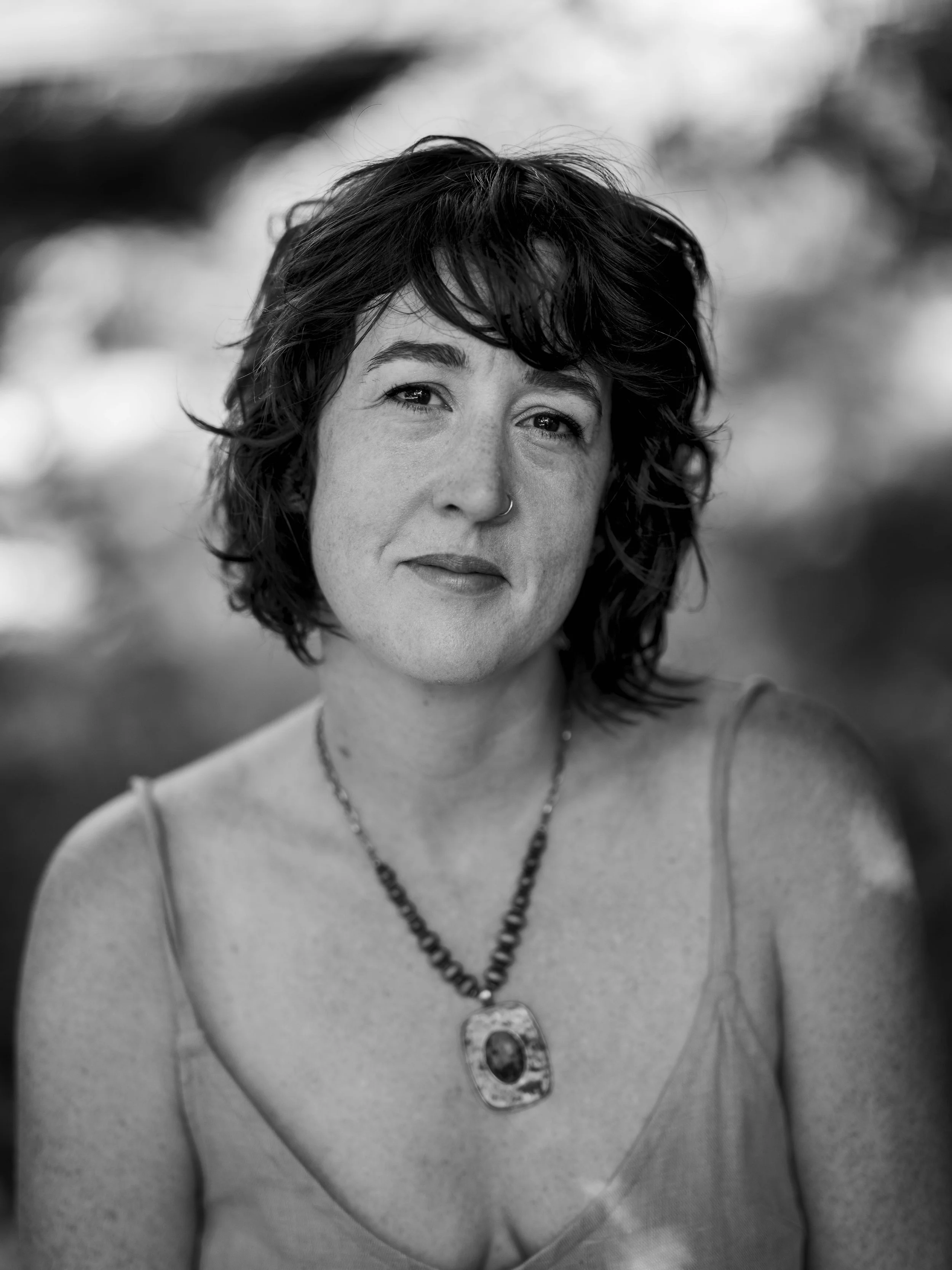 Black and white portrait of a woman with short, wavy hair, wearing a necklace and a sleeveless top, outdoors with blurred background.