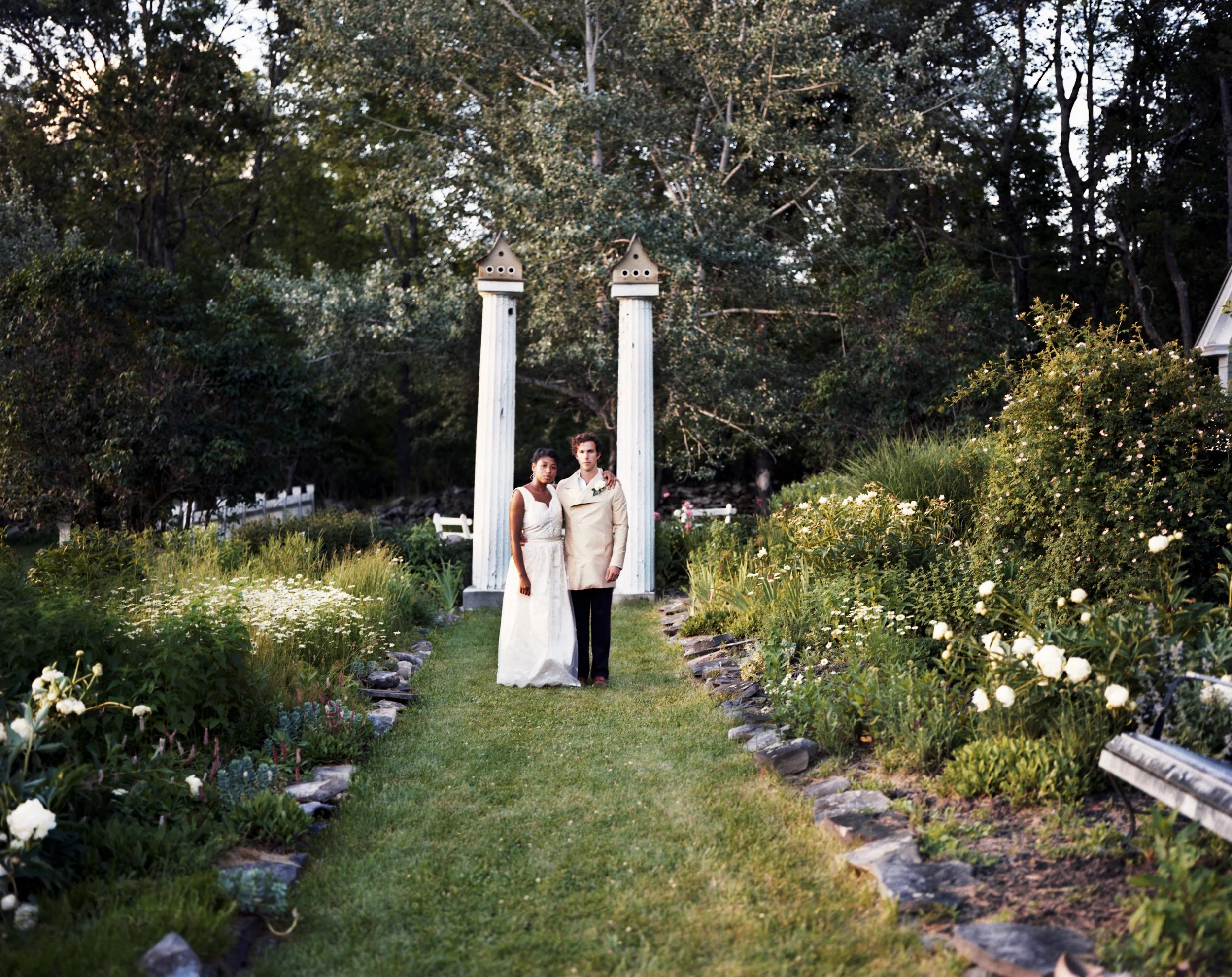 A wedding couple standing together on a garden path surrounded by flowers, with two white columns and trees in the background.