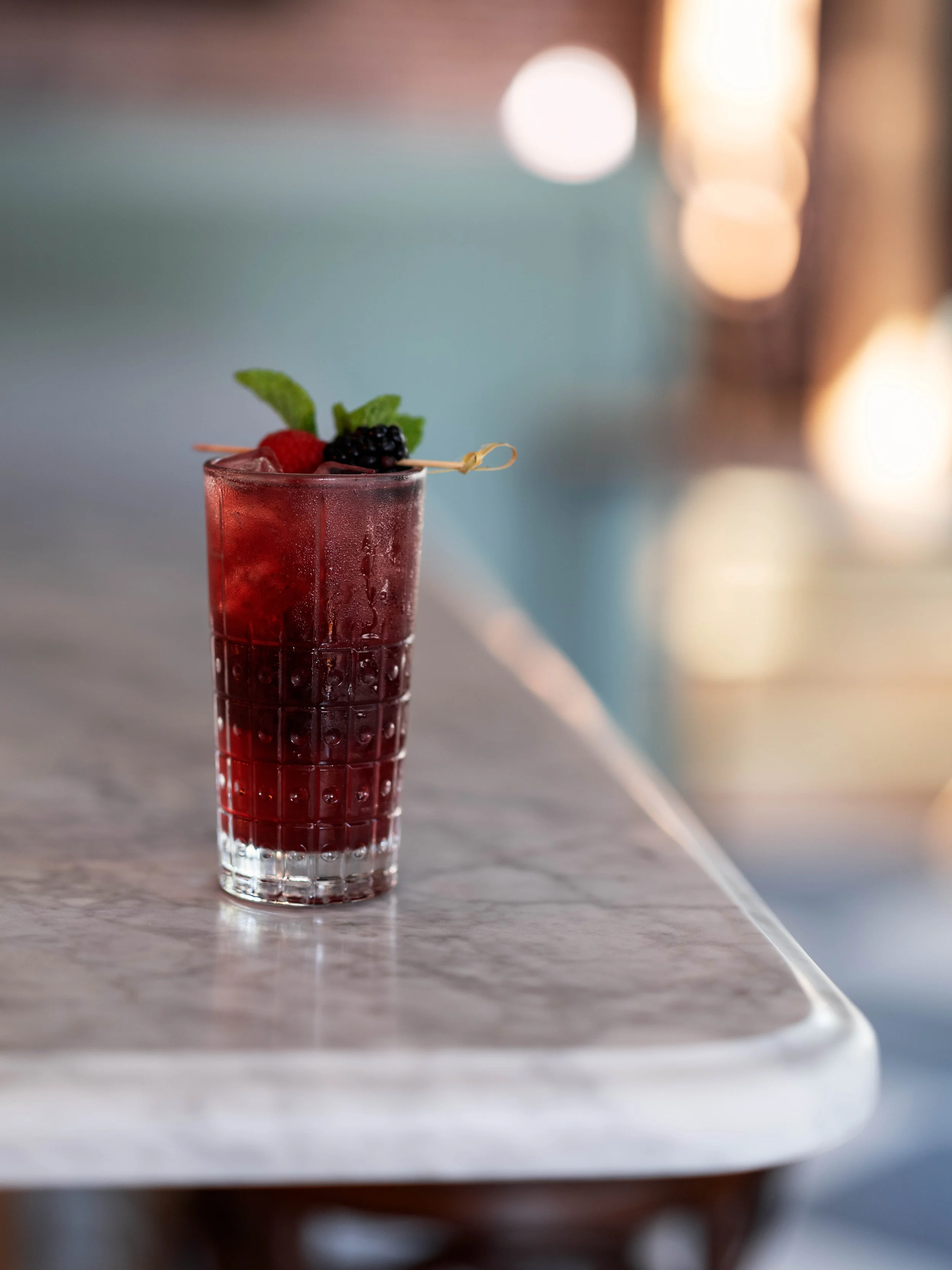 A tall glass of red berry cocktail garnished with fresh mint leaves, a raspberry, and a blackberry, on a marble table.