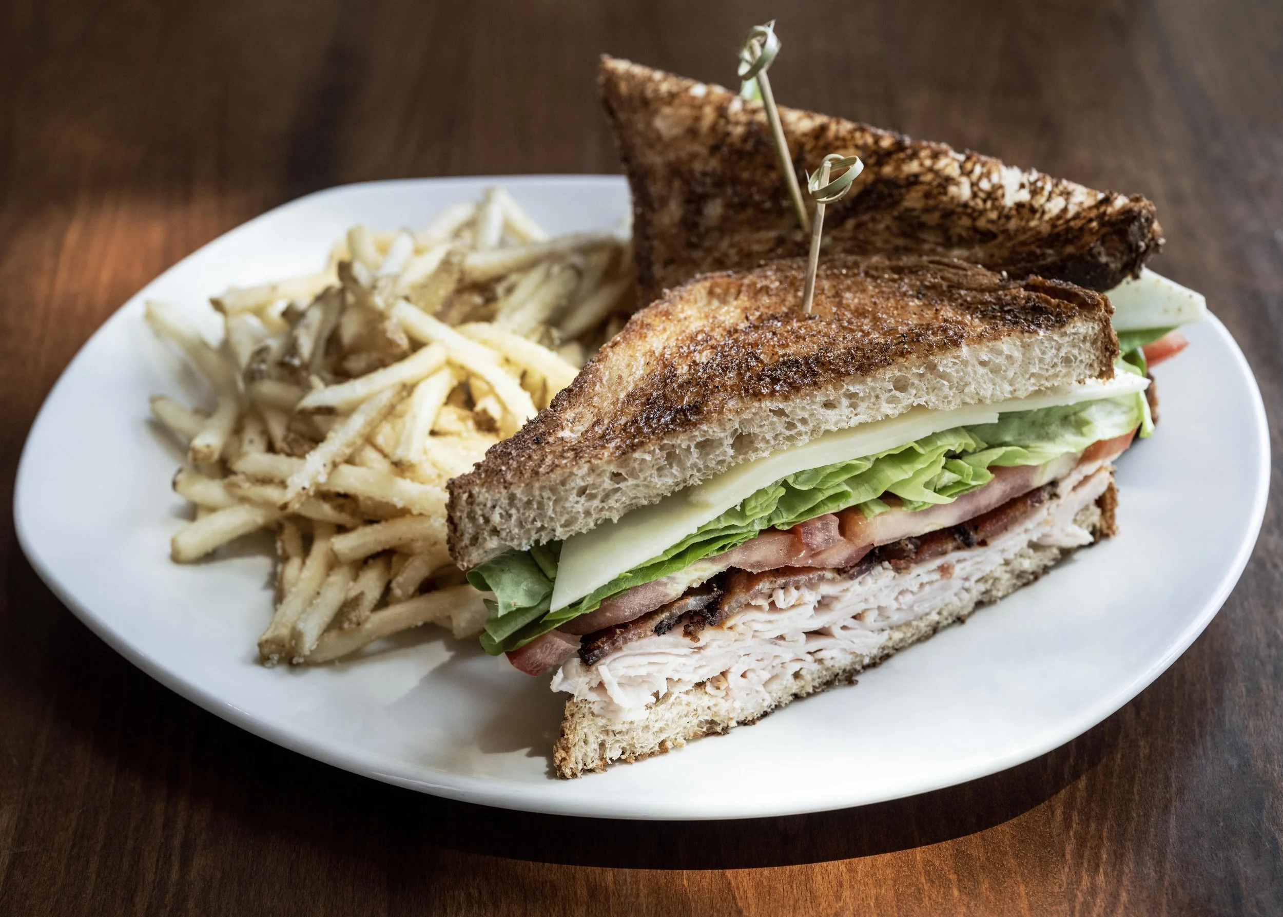 A plate with a large turkey club sandwich with lettuce, tomato, bacon, and cheese, served with a side of French fries, all on a white dish on a wooden table.