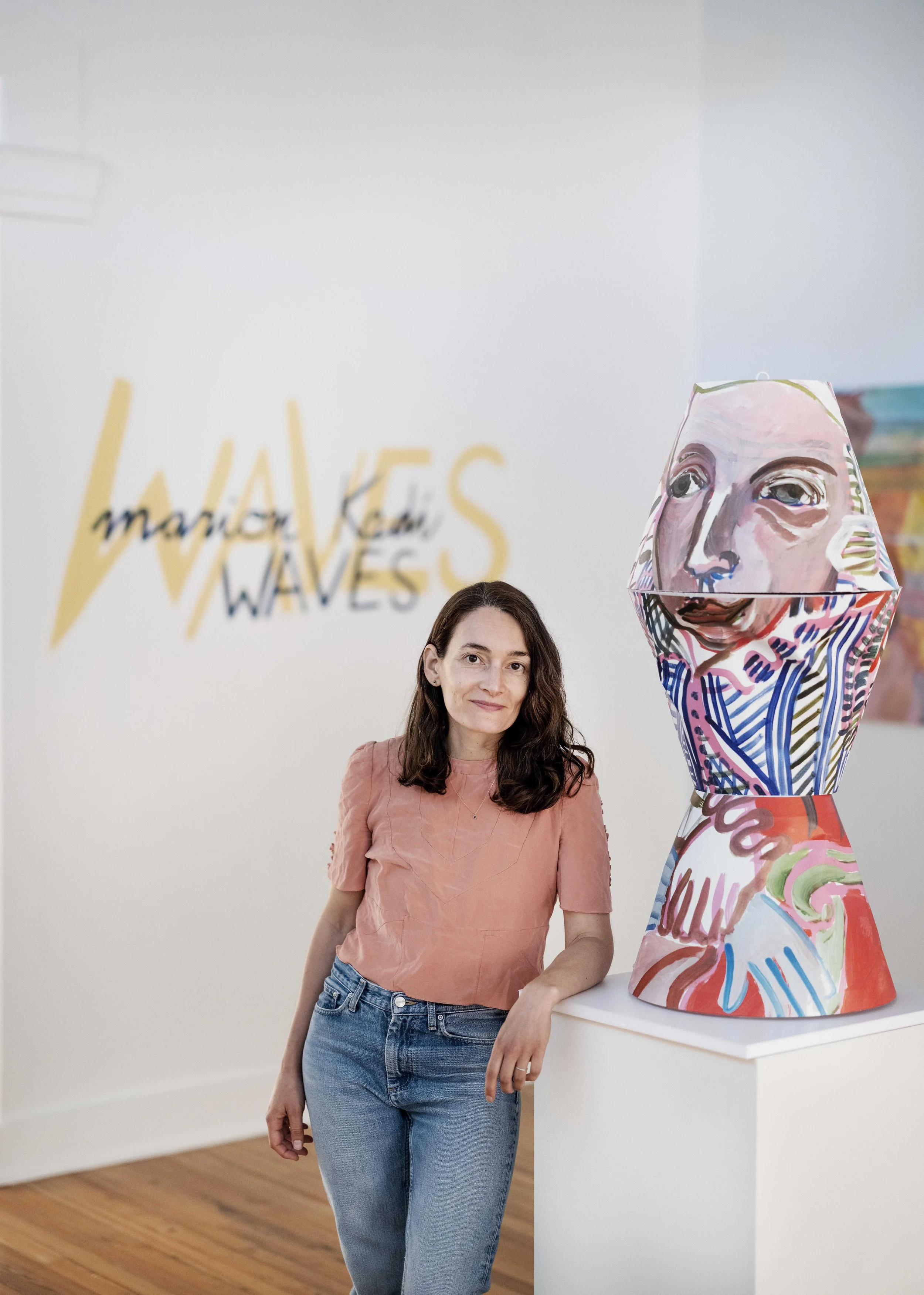 A woman with dark brown hair, wearing a peach-colored shirt and blue jeans, standing next to a colorful abstract sculpture with a human face painted on it. In the background, there is a blurred wall with the artwork title 'WAVES' and the artist's nam