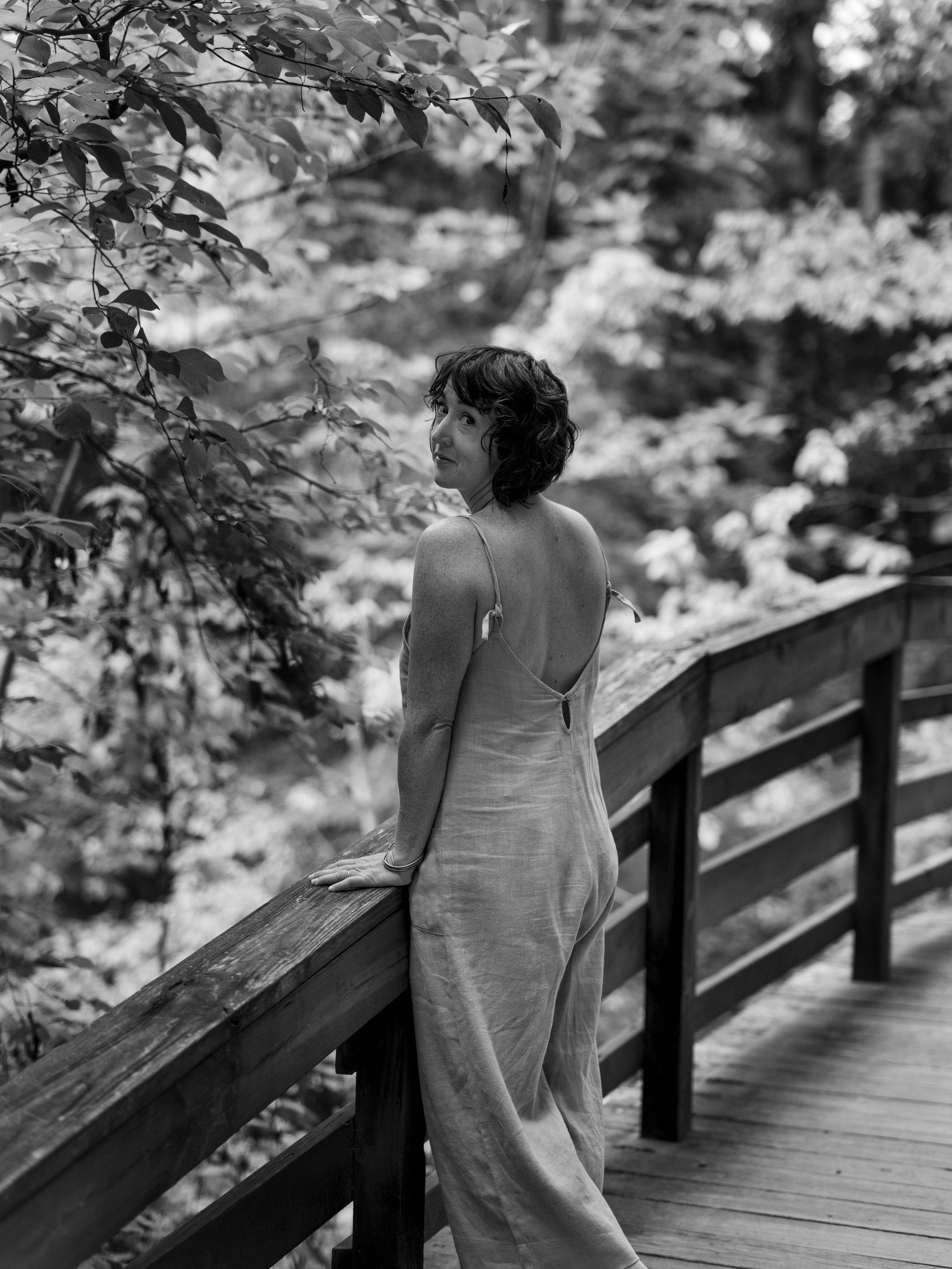 A woman with short curly hair wearing a backless dress, standing on a wooden bridge in a natural forest setting, looking over her shoulder.