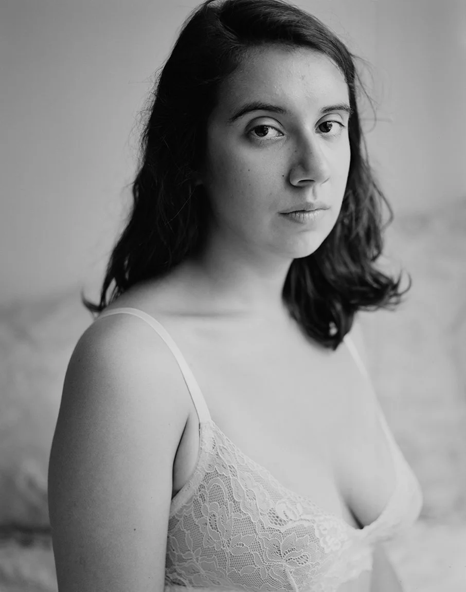 Black and white photo of a young woman with shoulder-length dark hair, wearing a lace camisole, looking directly at the camera with a serious expression.