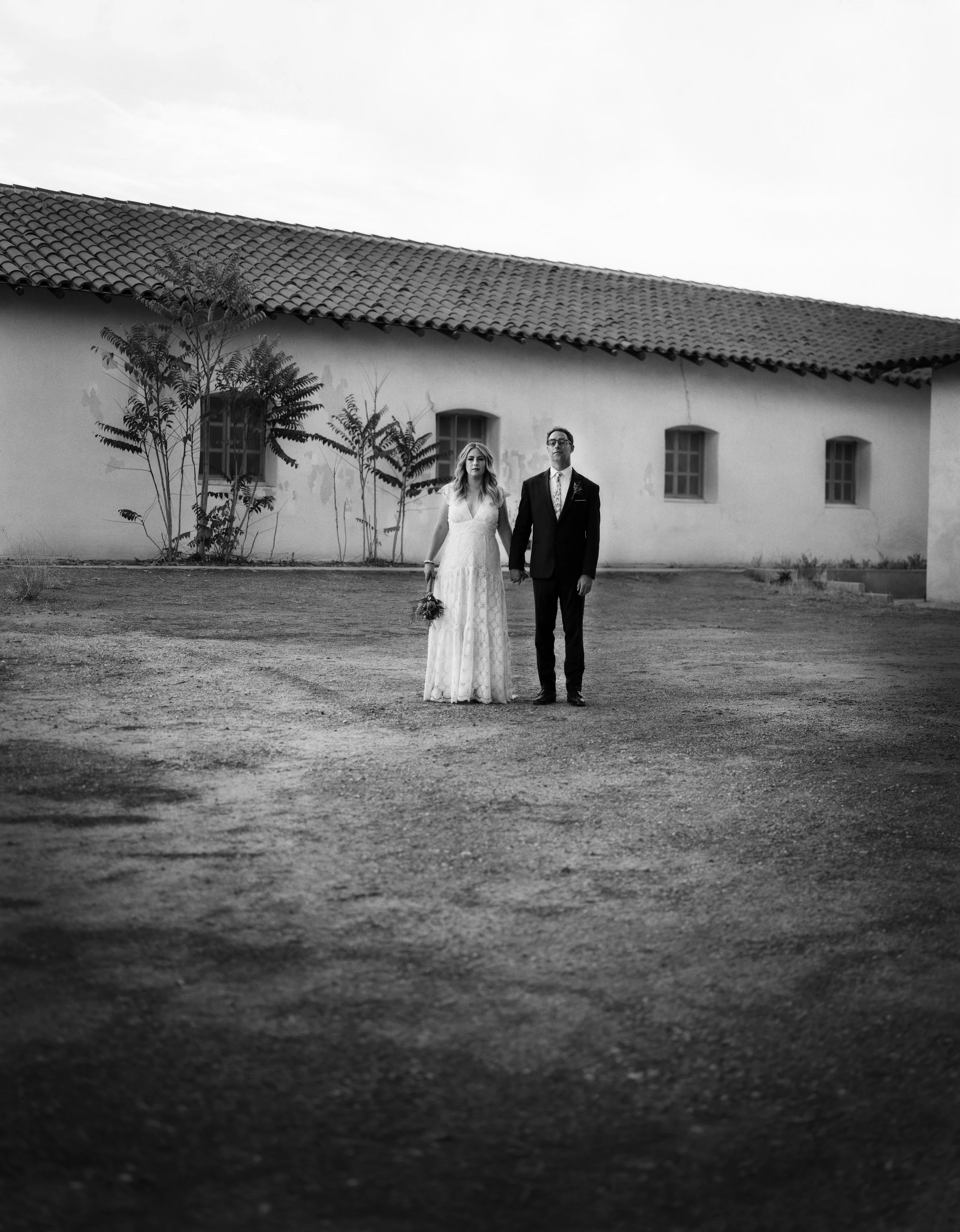 Black and white photo of a bride and groom standing hand in hand outdoors in front of a building with small windows and a tiled roof, with two small trees along the wall.