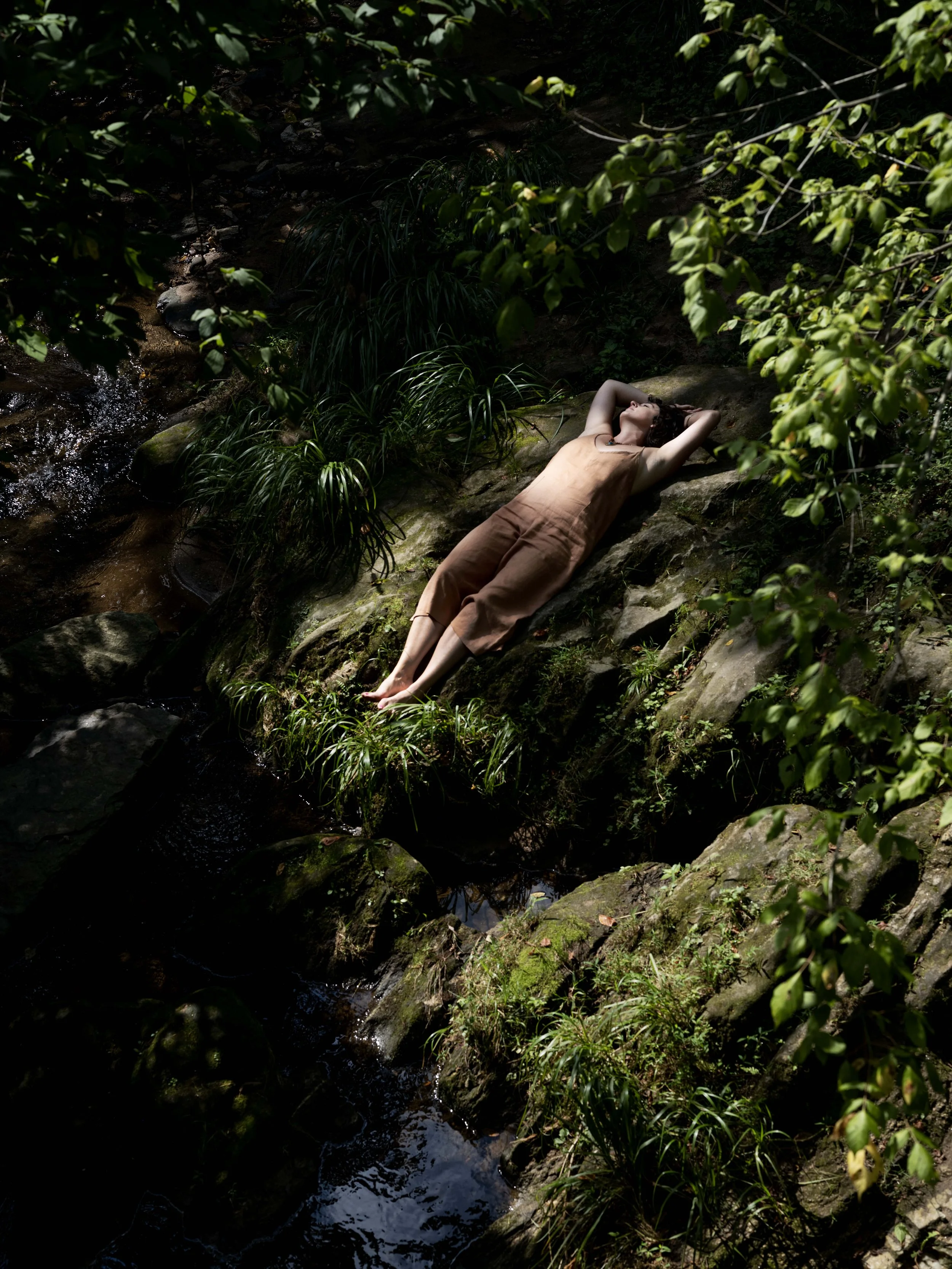 Woman lying on mossy rocks by a stream in a forest, with her eyes closed and arms resting behind her head, surrounded by greenery.