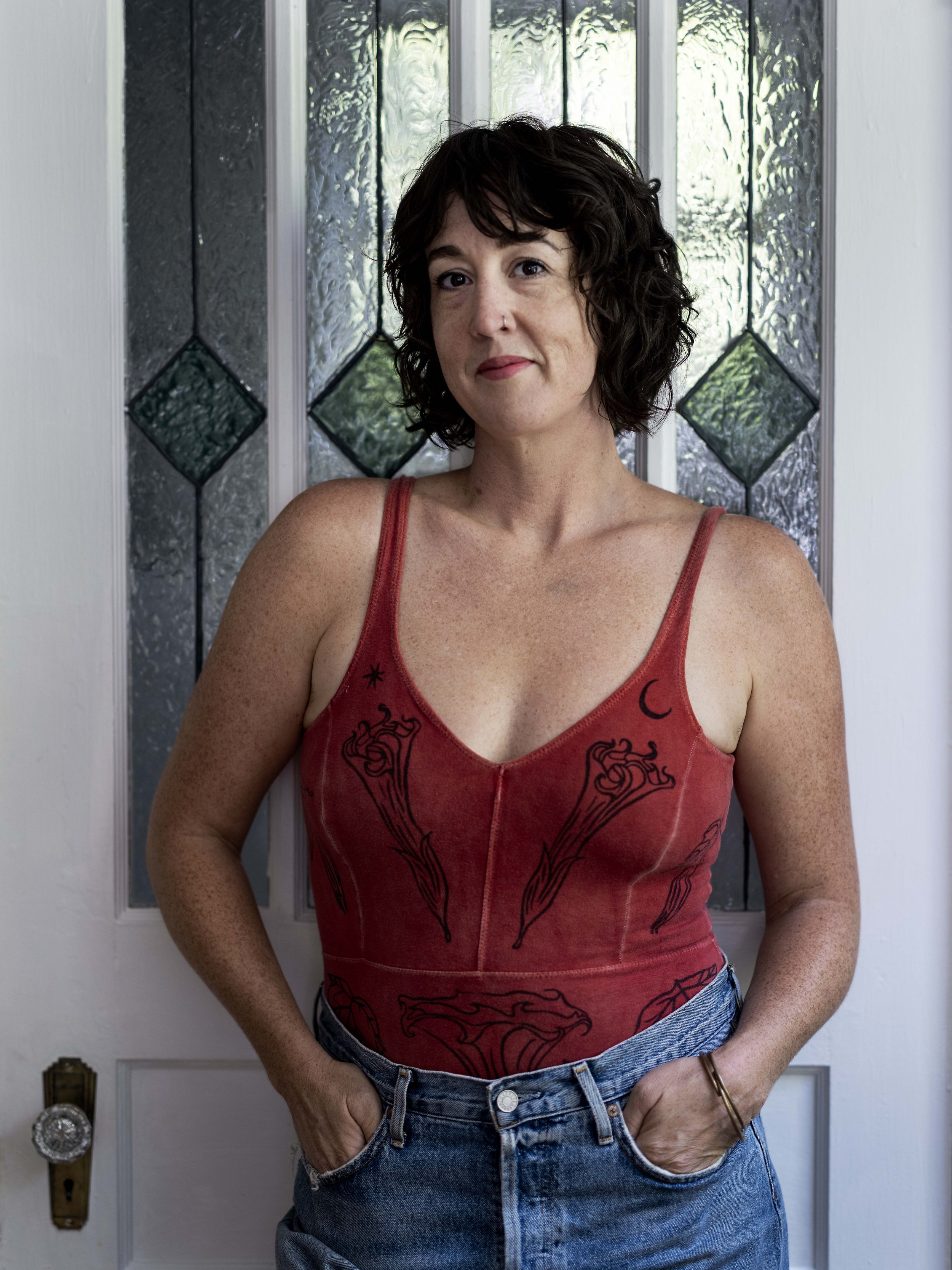 A woman with short, curly dark hair standing in front of a stained glass door, wearing a red tank top with black designs and blue jeans, with hands in her pockets.