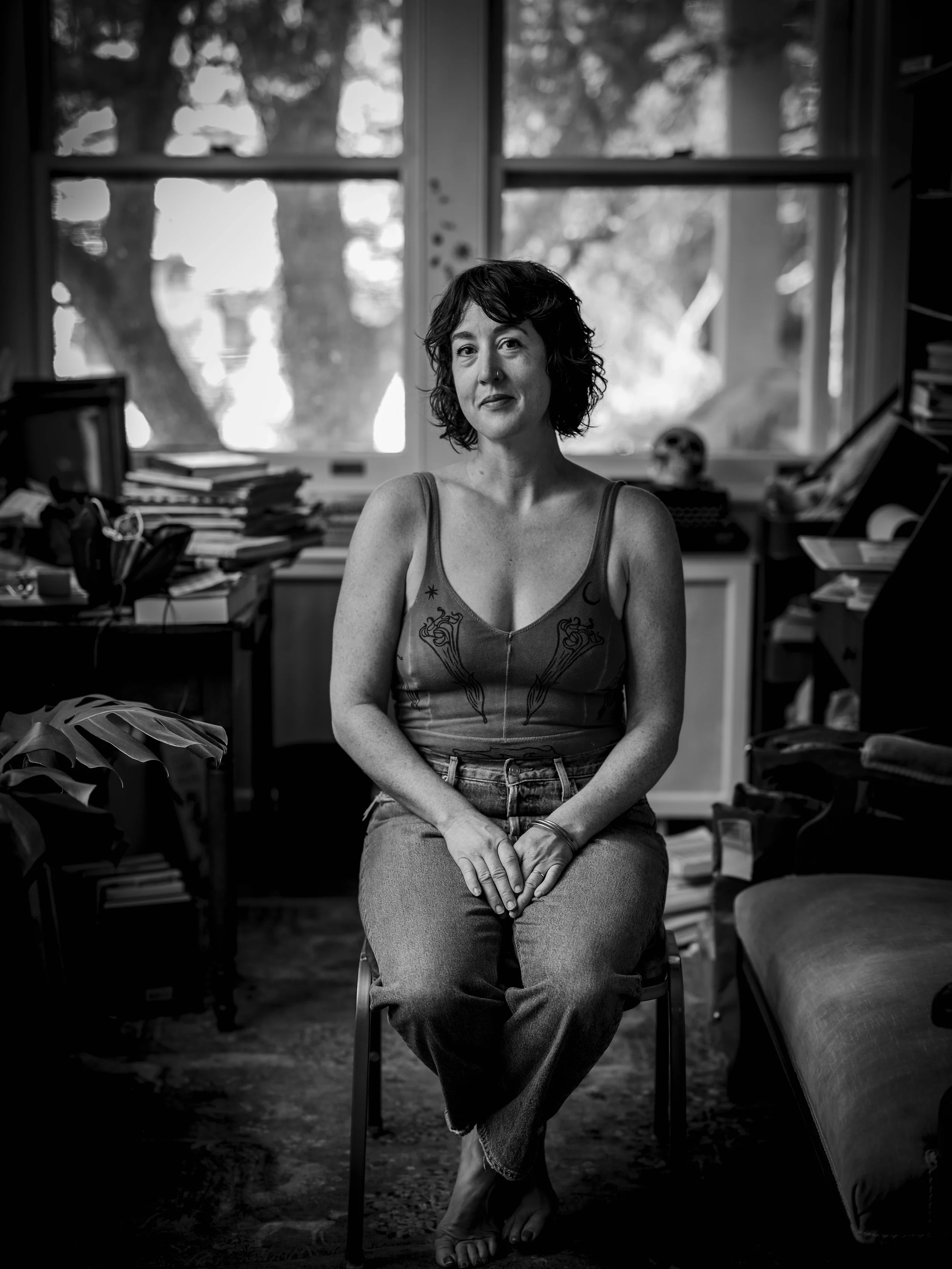 A woman with curly hair sits on a chair in a cluttered room with large windows, stacks of books, and a small skull on a desk, black and white photography.