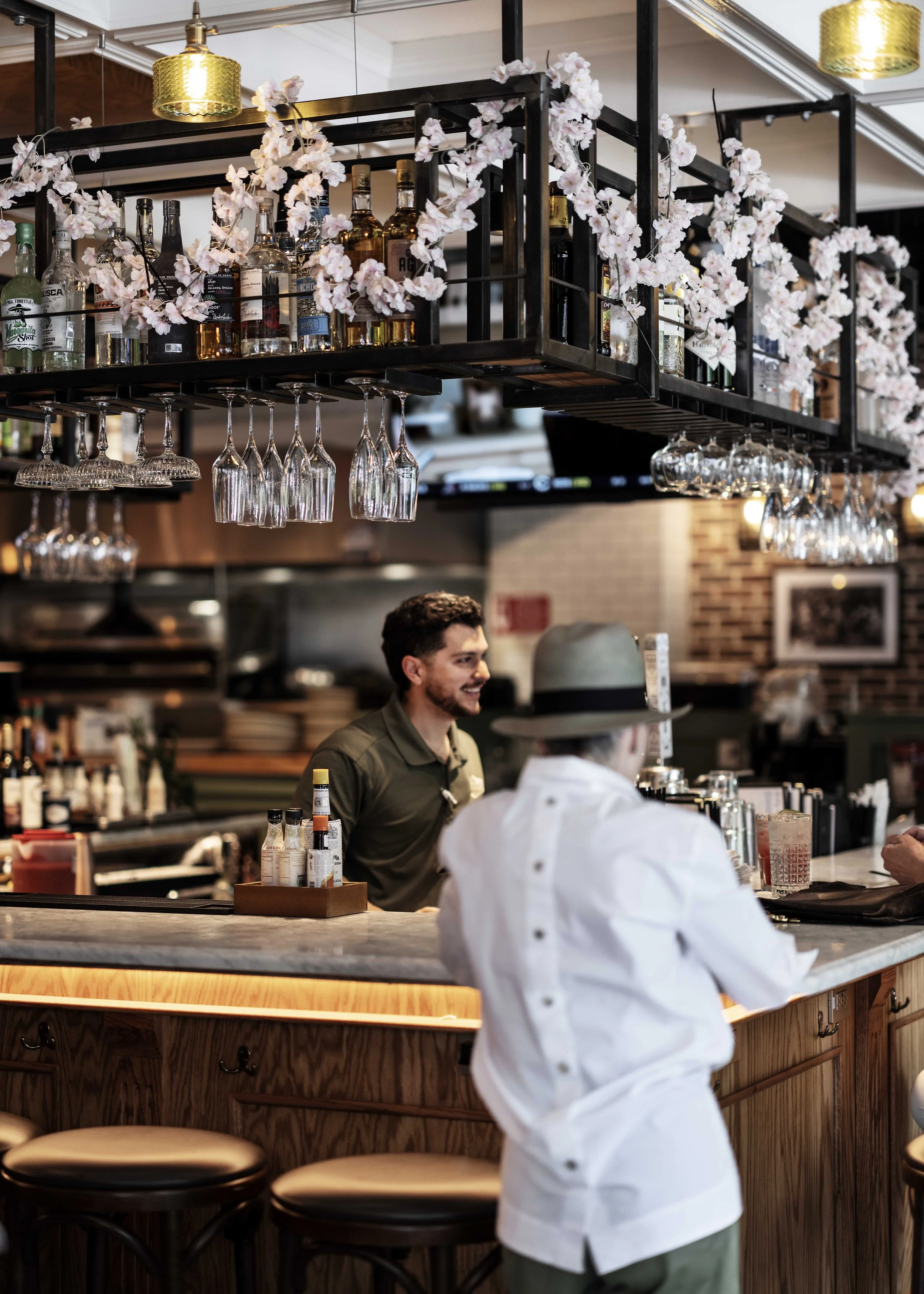 A bartender and a guest engaged in conversation at a bar with a marble countertop and wooden paneling. The bar is decorated with hanging glassware, flower garlands, and bottles of liquor. The background shows kitchen shelves and brick walls.