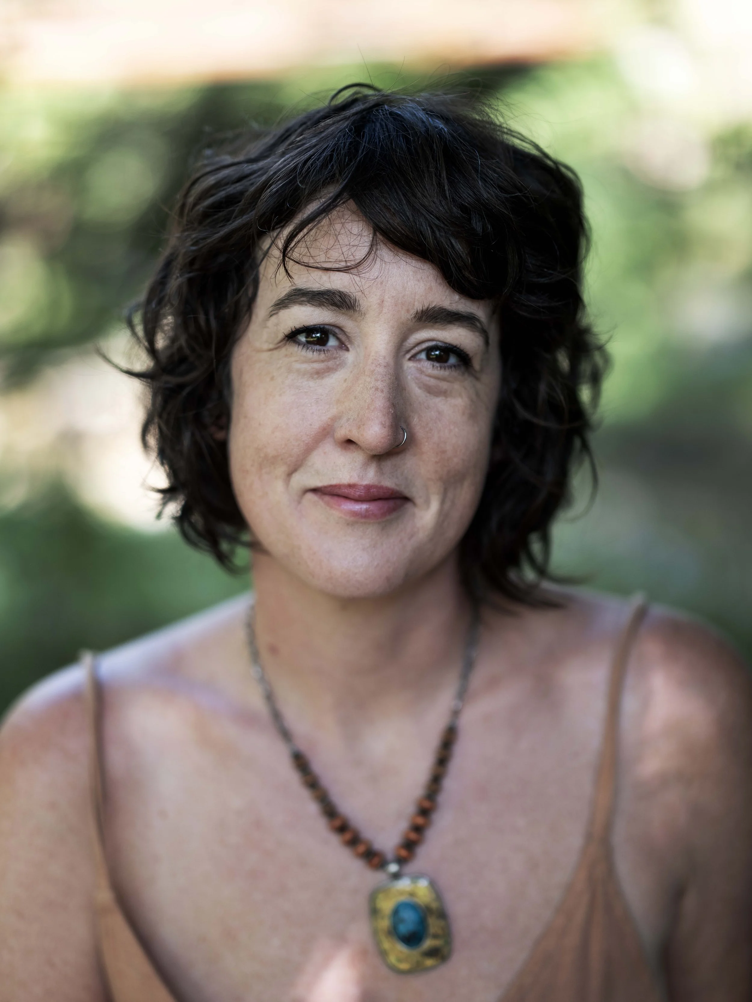 Close-up of a woman with dark curly hair, wearing a tan spaghetti strap top and a necklace with a large pendant. She has a slight smile, light makeup, and a nose ring, with a blurred green natural outdoor background.