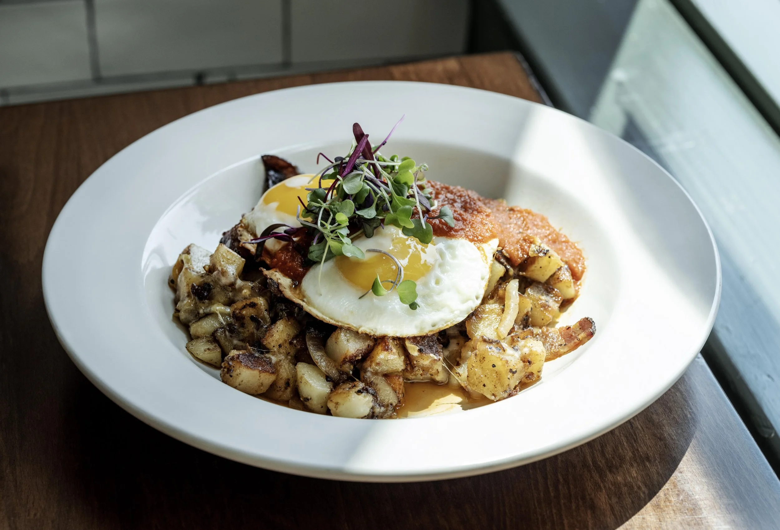 Dish of roasted potatoes topped with two sunny side up eggs, tomato sauce, microgreens, and a poached egg on top, served on a white plate near a window.