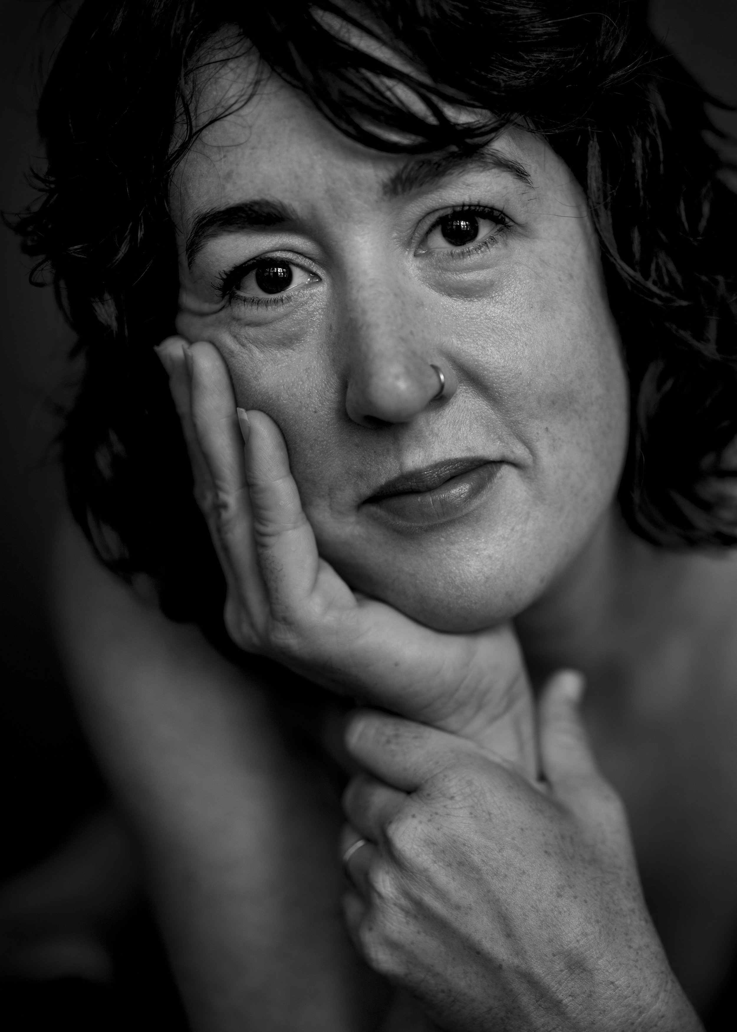 Black and white close-up portrait of a woman with curly hair, a nose ring, and freckles, resting her face on her hand.