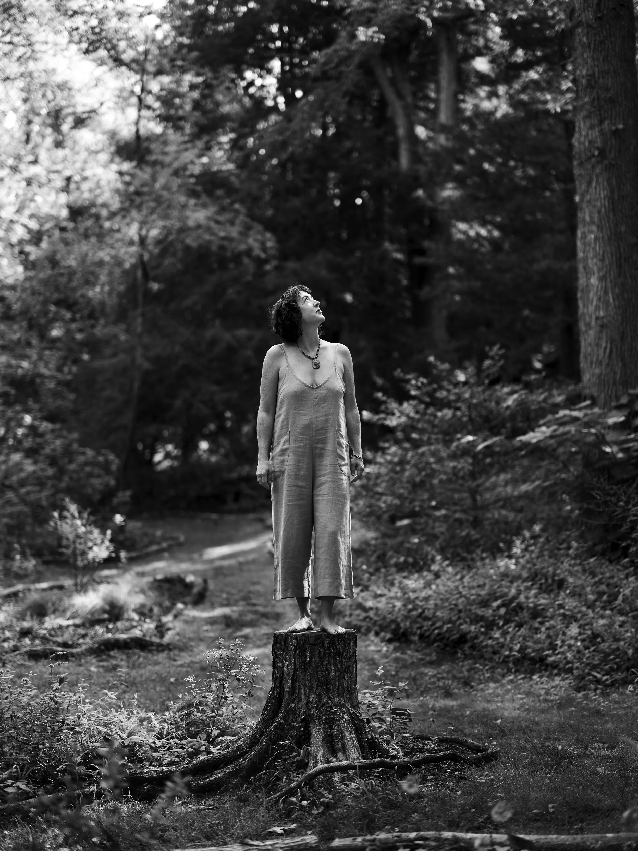 A woman in a jumpsuit standing barefoot on a tree stump in a forest, looking upward.