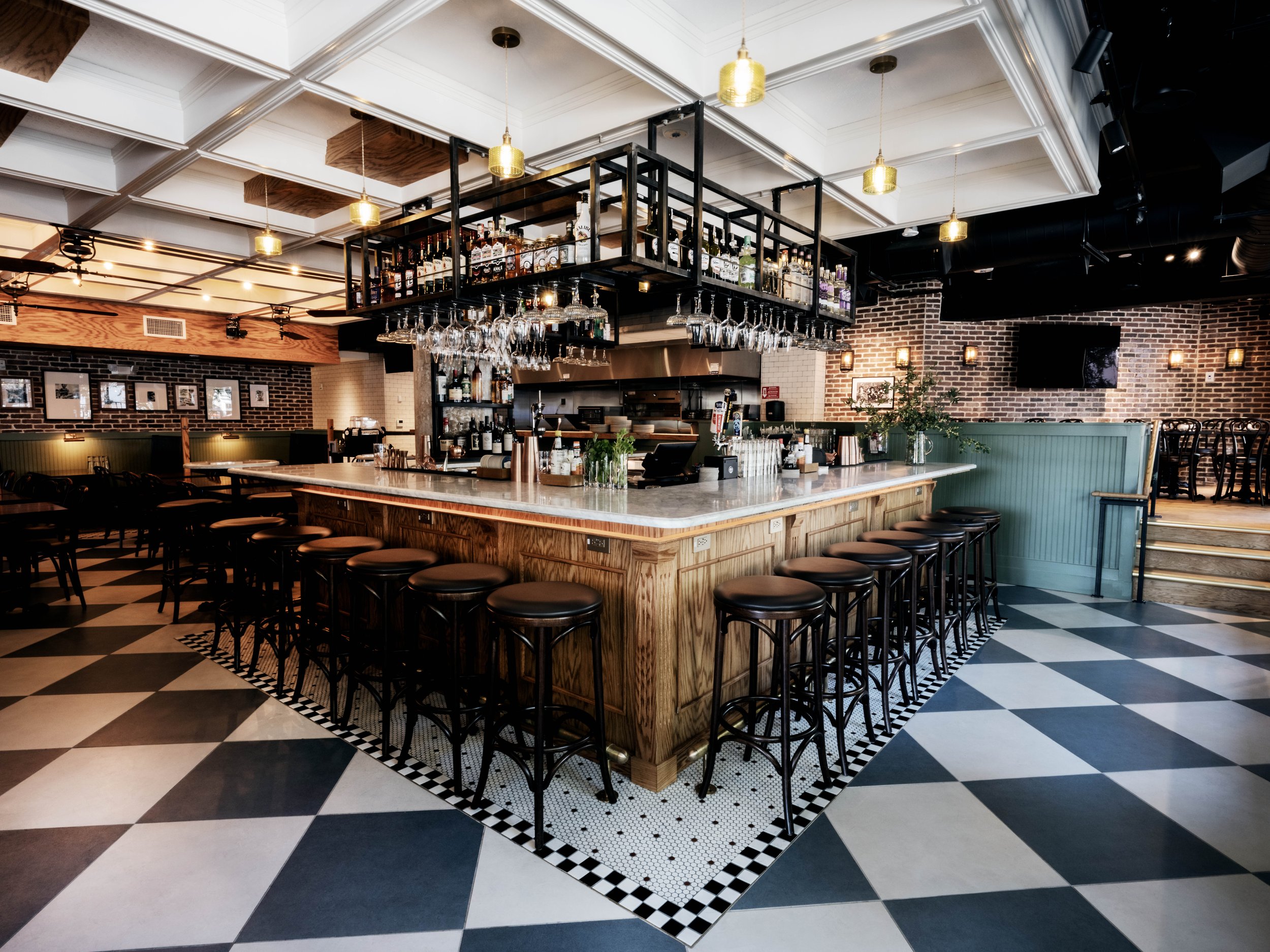 Empty bar with wooden countertop and black barstools, hanging wine glasses, liquor bottles on shelves, checkered black and white floor, and brick walls in a stylish restaurant interior.