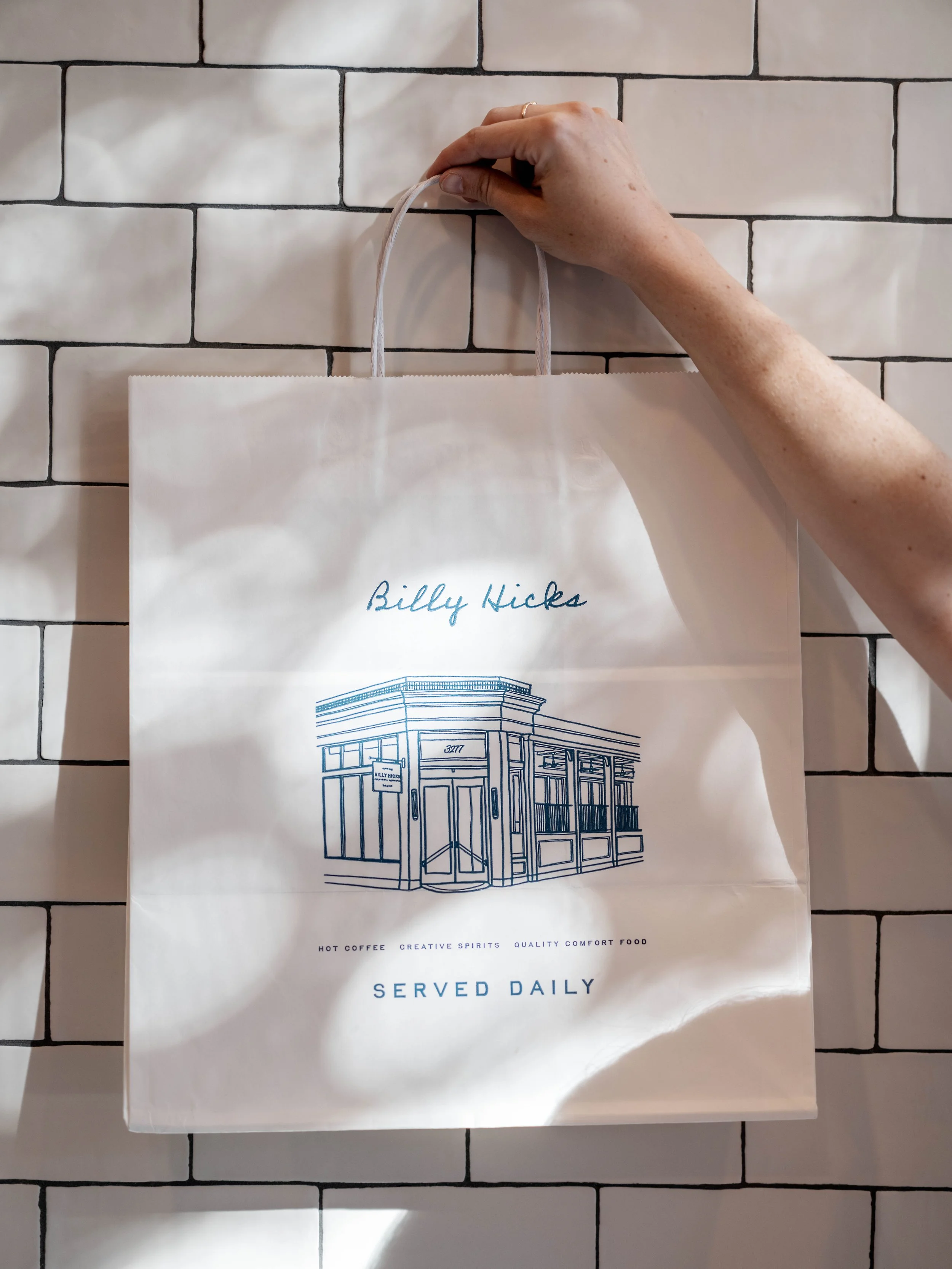 A white paper shopping bag with the name 'Billy Hicks' and an illustration of a building exterior, held up against a white tiled wall by a hand.