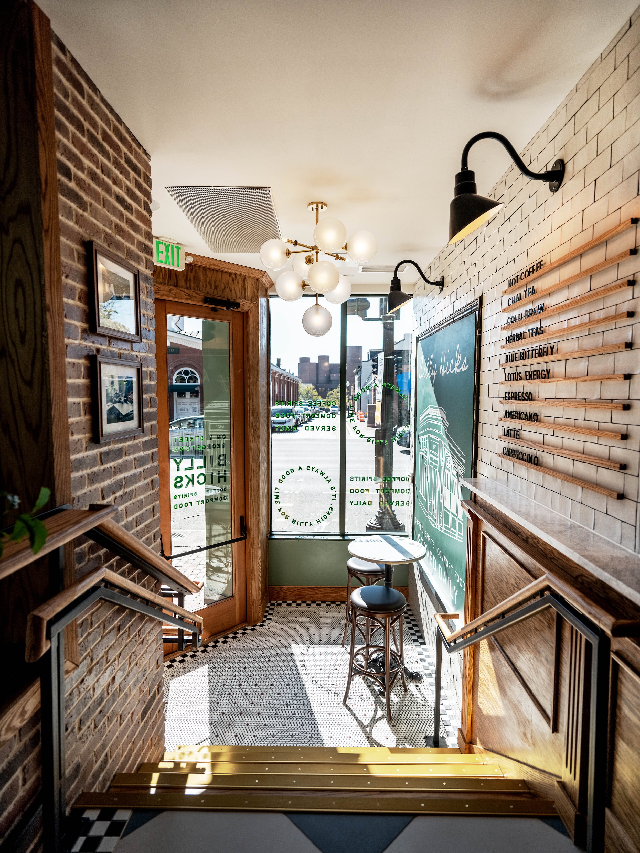 Interior of a cozy coffee shop with a brick and white tile wall, black wall-mounted lamps, a white round table, two black stools, and a menu written on wood slats on the wall. Sunlight streams through the glass door and window, illuminating the tiled