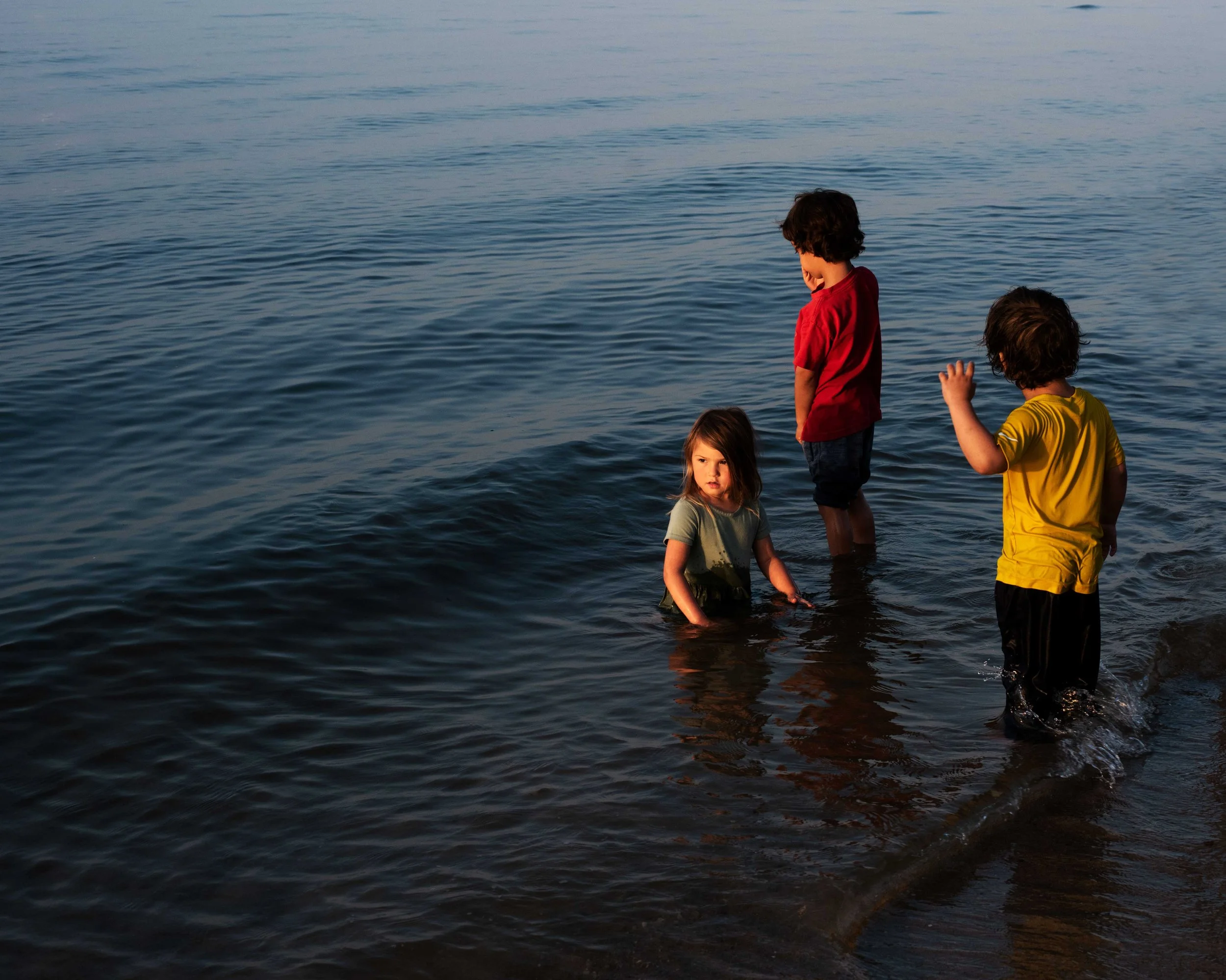 Three children standing in shallow water at the beach during sunset.