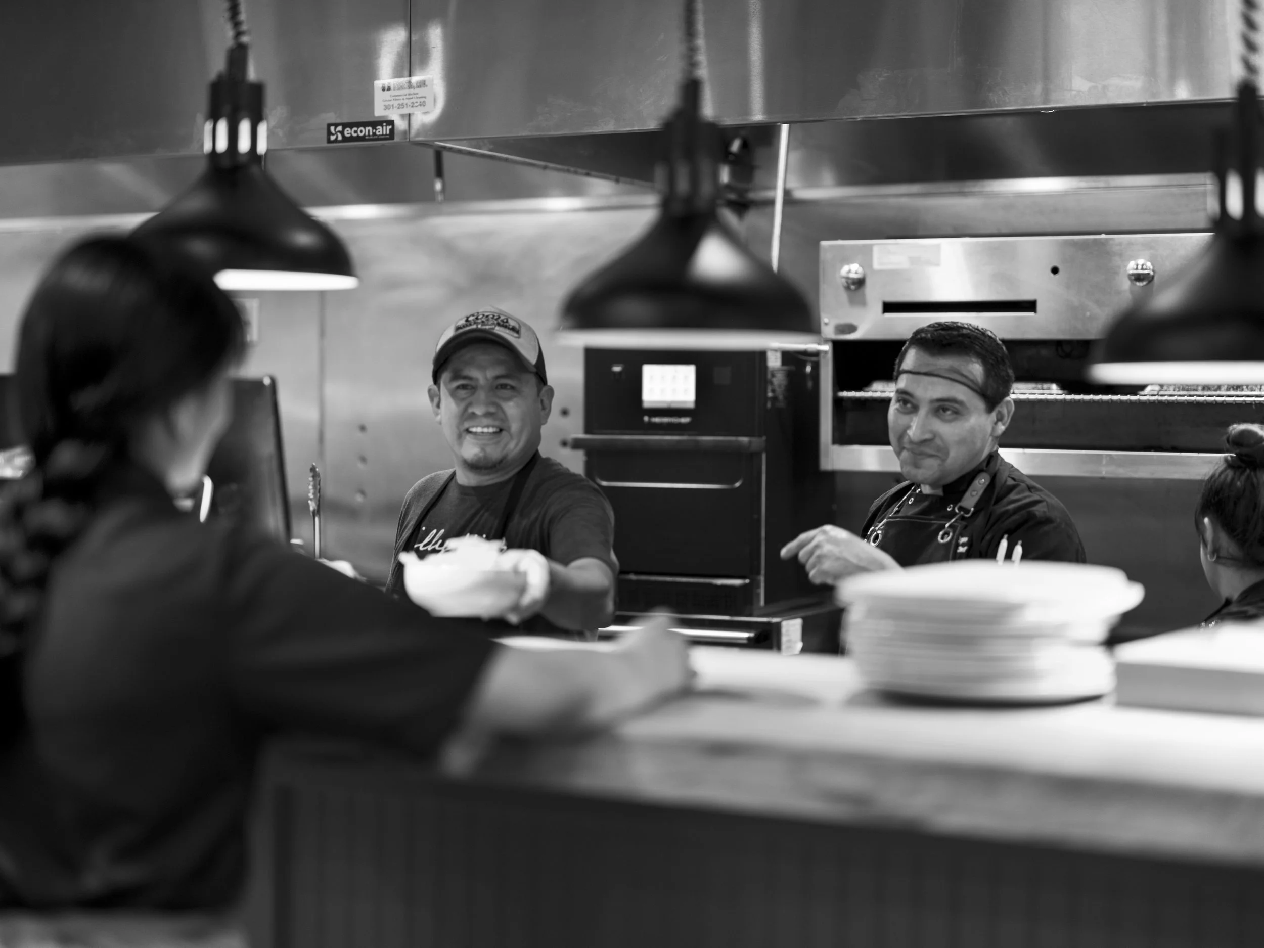 Black and white photo of restaurant kitchen staff interacting with a customer at the counter. Two cooks or chefs are smiling, one handing something to the customer, while the other looks on. Plates are stacked on the counter in the foreground.