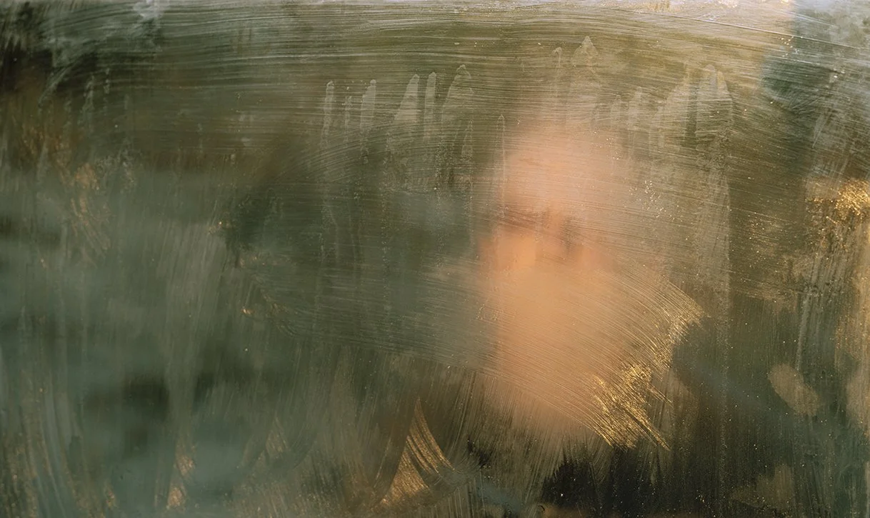 A person's face is partially visible through a foggy or dirty glass surface, with blurred features and smudges on the glass.